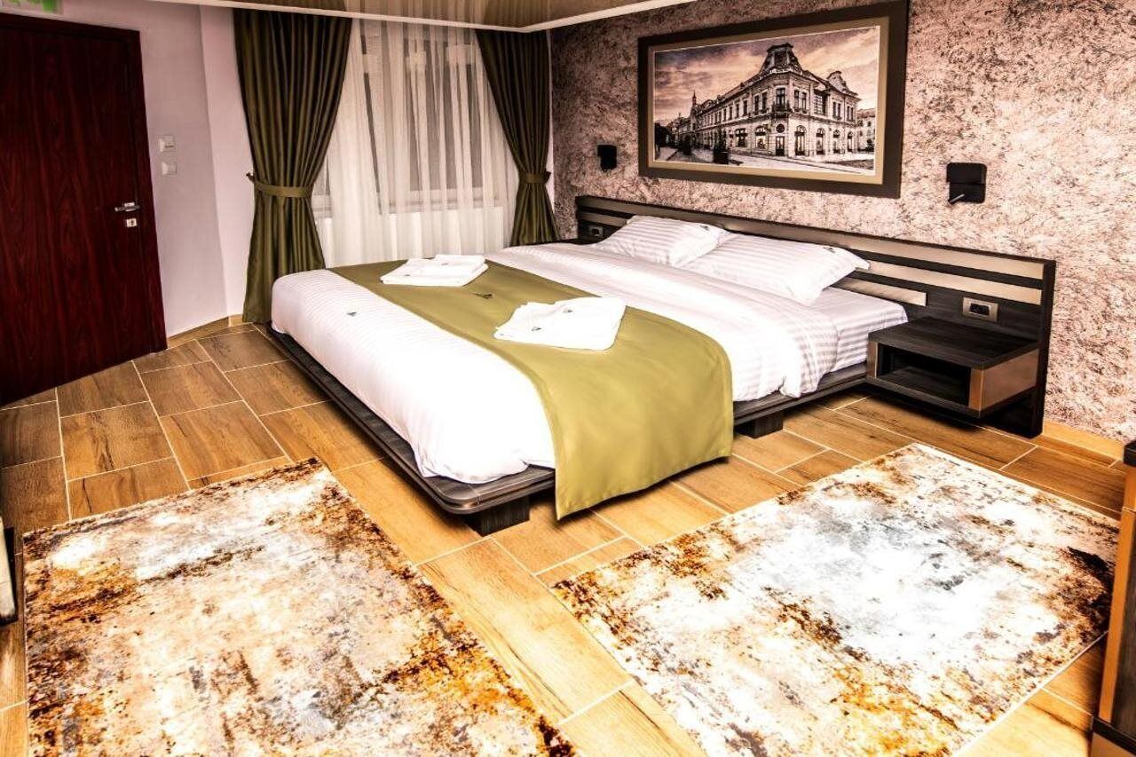 Een hotelkamer met een groot bed, een nachtkastje, een aan de muur bevestigde televisie en een lijst met een afbeelding van een gebouw.