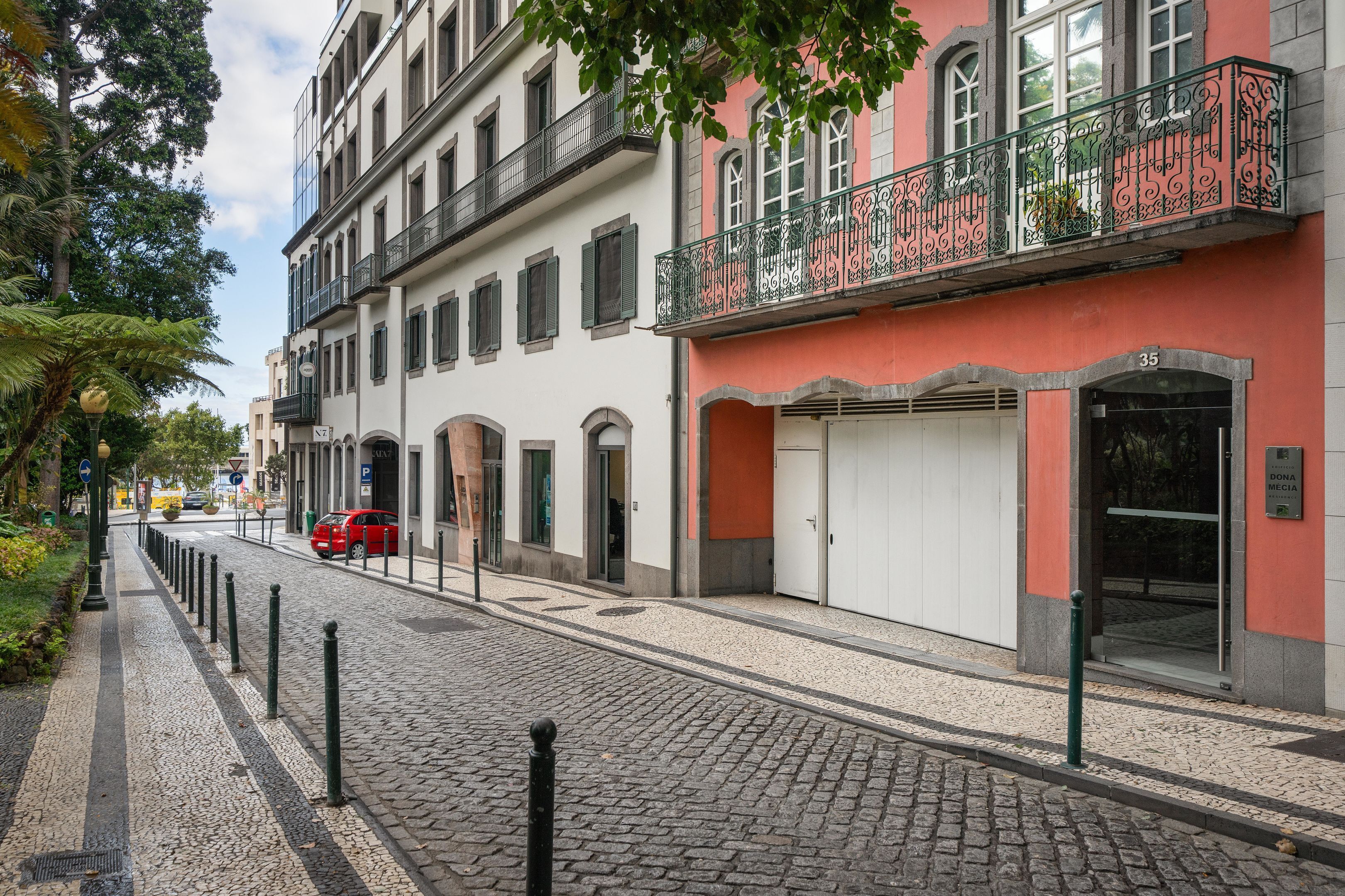 A cobblestone street with buildings, a red car, and a sign for 'Dona Media'.