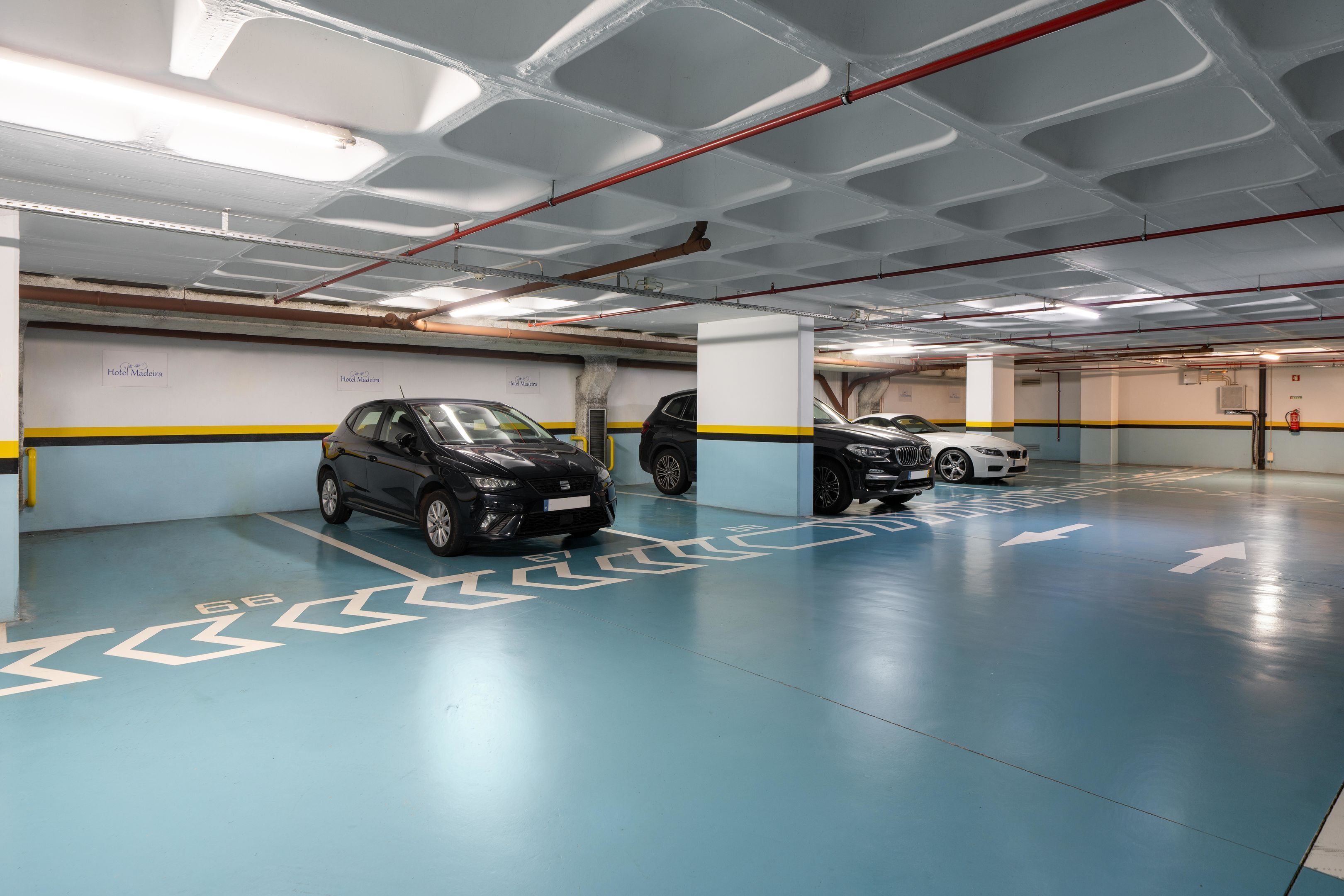 Un aparcamiento de varios niveles con flechas direccionales claras en el suelo, varios coches estacionados y un cartel que indica 'Hotel Madeira'.