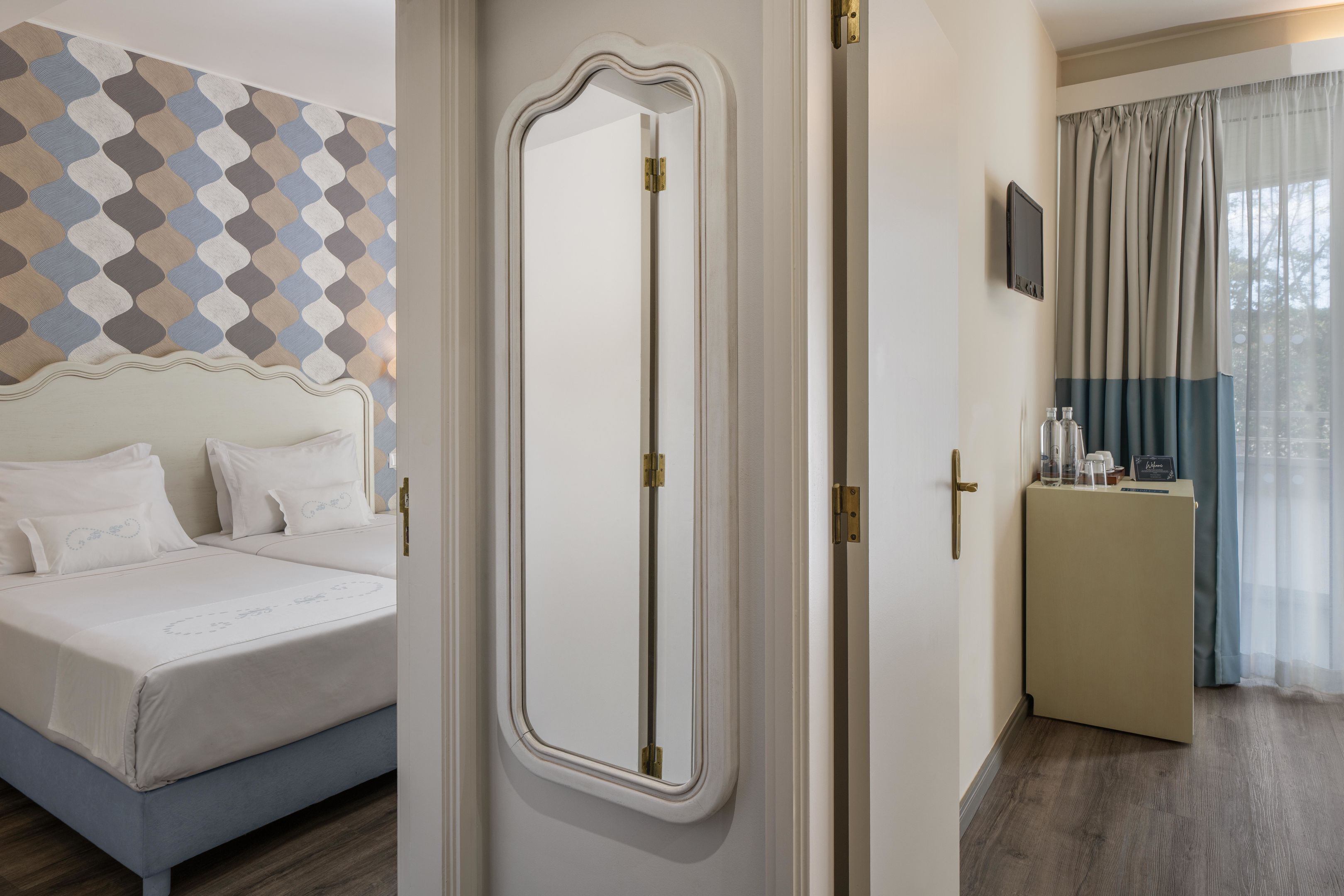 A hotel room with a bed, a mirror, a small table with items, and a view through a window with curtains.