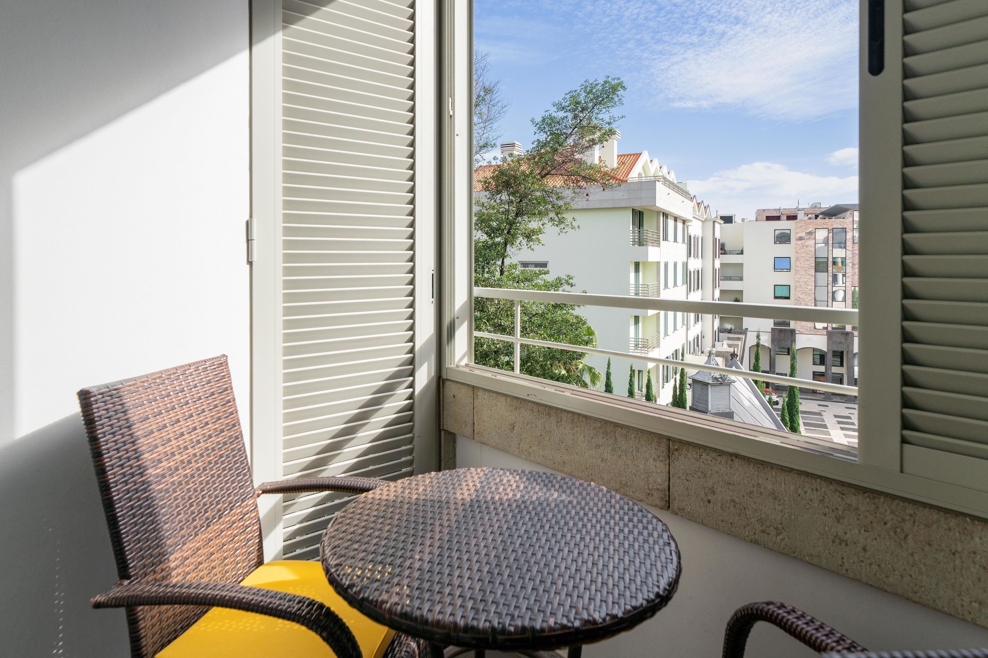 Een balkon met rieten stoelen, een klein rond tafeltje en uitzicht op gebouwen door het raam.