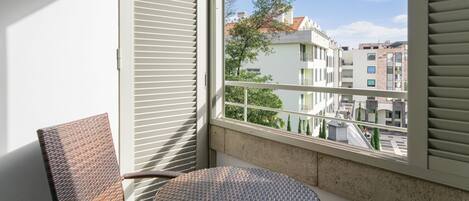 Een balkon met rieten stoelen, een klein rond tafeltje en uitzicht op gebouwen door het raam.
