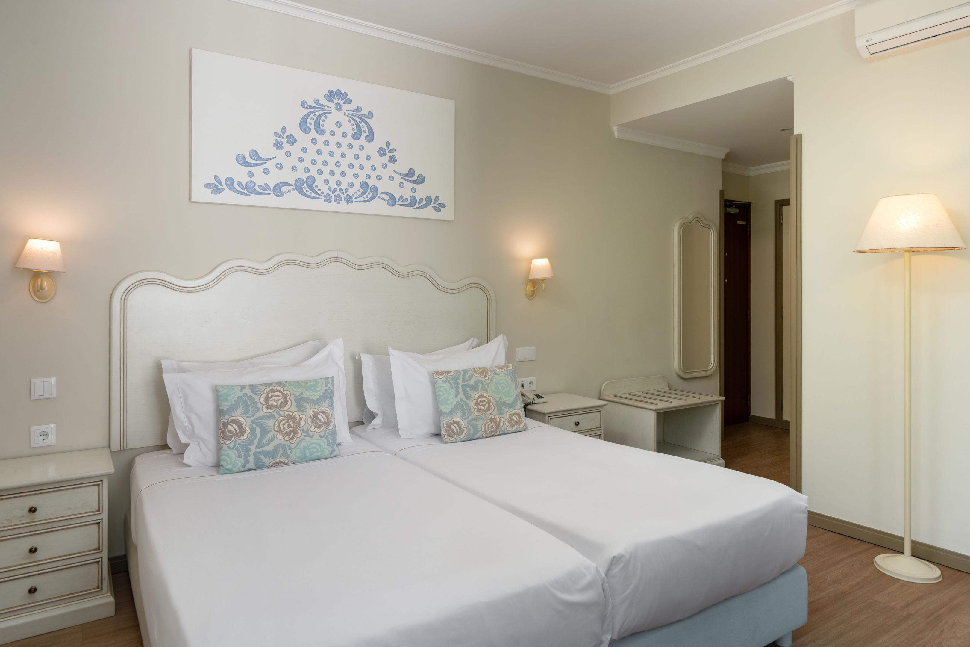 A double bed with white bedding and floral-patterned pillows, bedside tables with lamps, a wall-mounted artwork, and a mirror.
