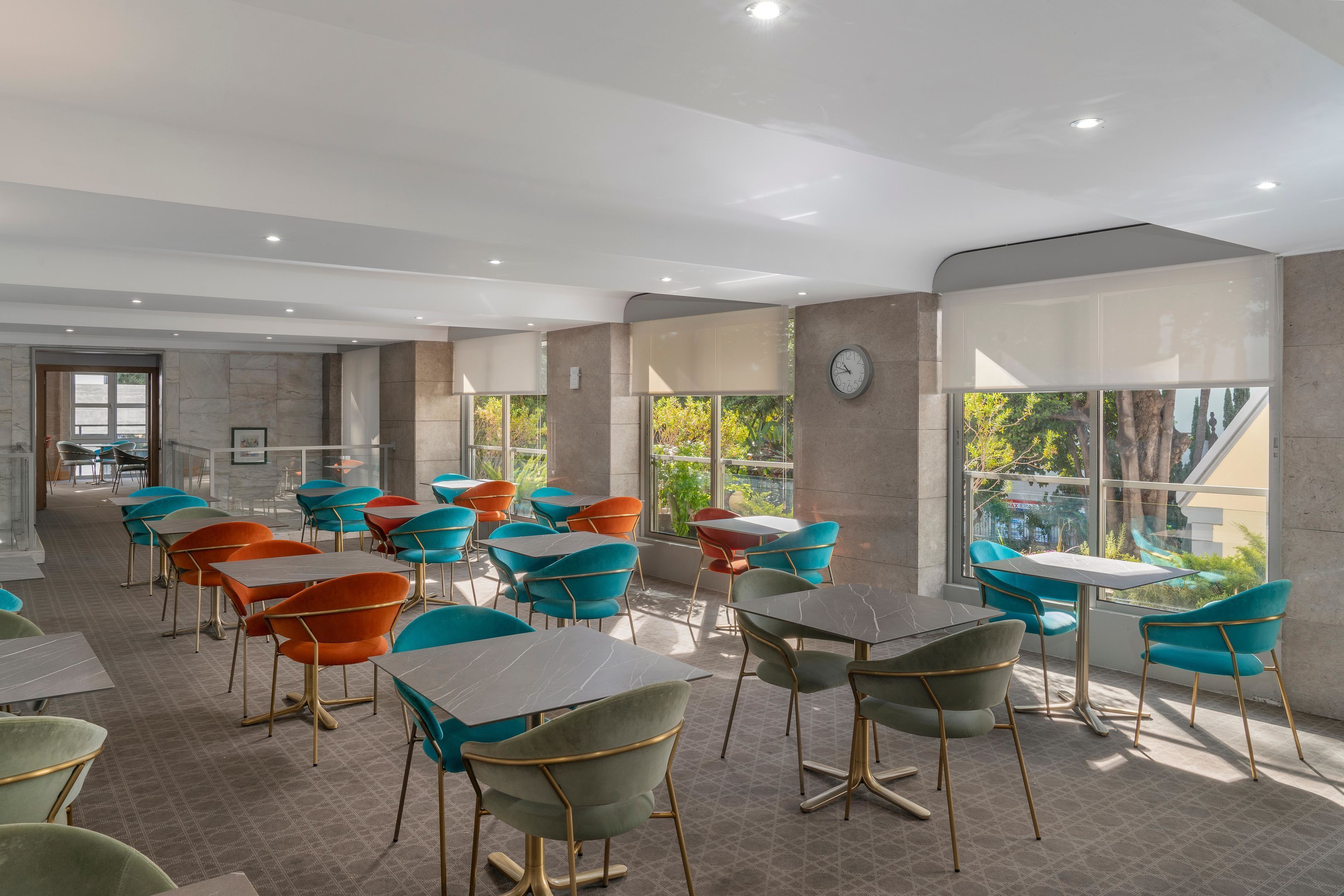 A modern dining area with colorful chairs and tables, large windows, and a clock on the wall.