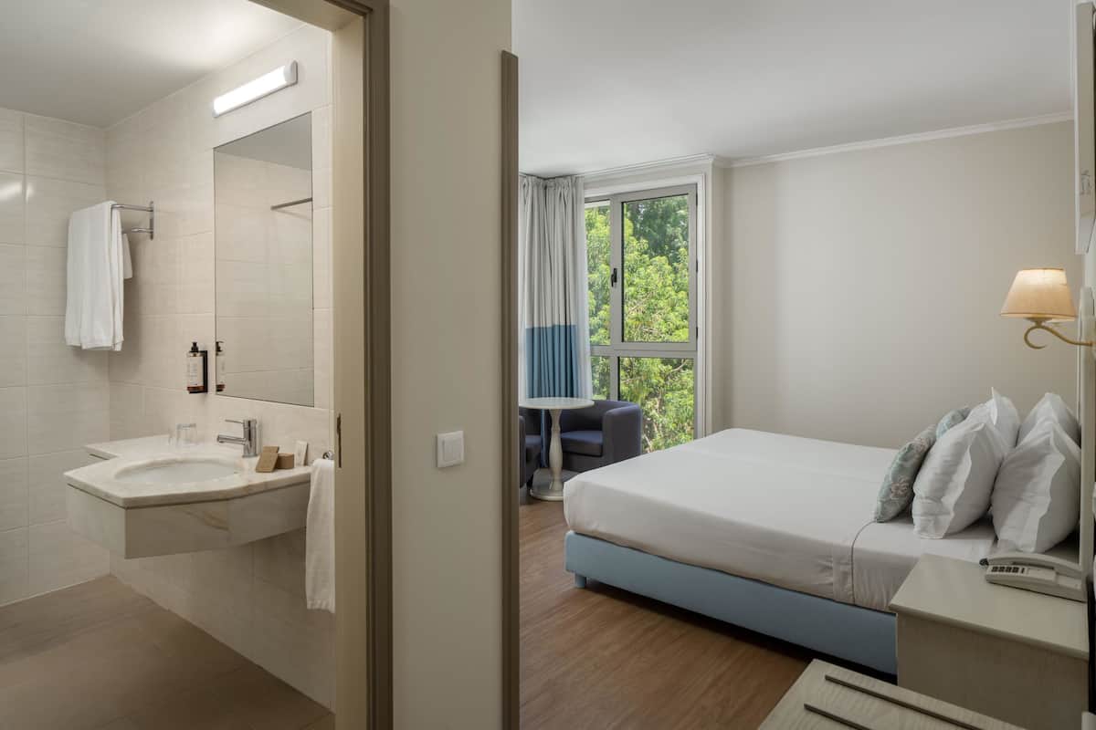 Ein modernes Hotelzimmer mit Bad, Bett, Nachttisch und Fenster mit Blick auf Grünflächen.