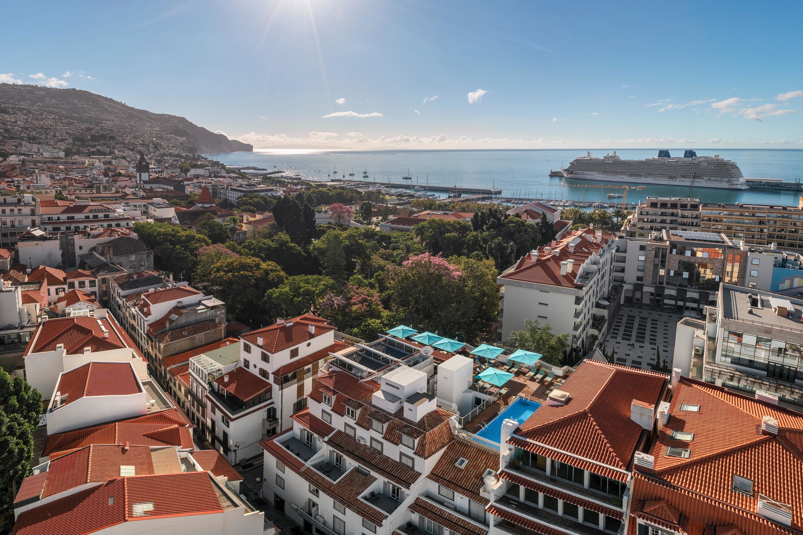 Un paisaje urbano costero con un gran barco crucero amarrado en el puerto, rodeado de edificios residenciales con techos de tejas rojas y una vista despejada del mar.