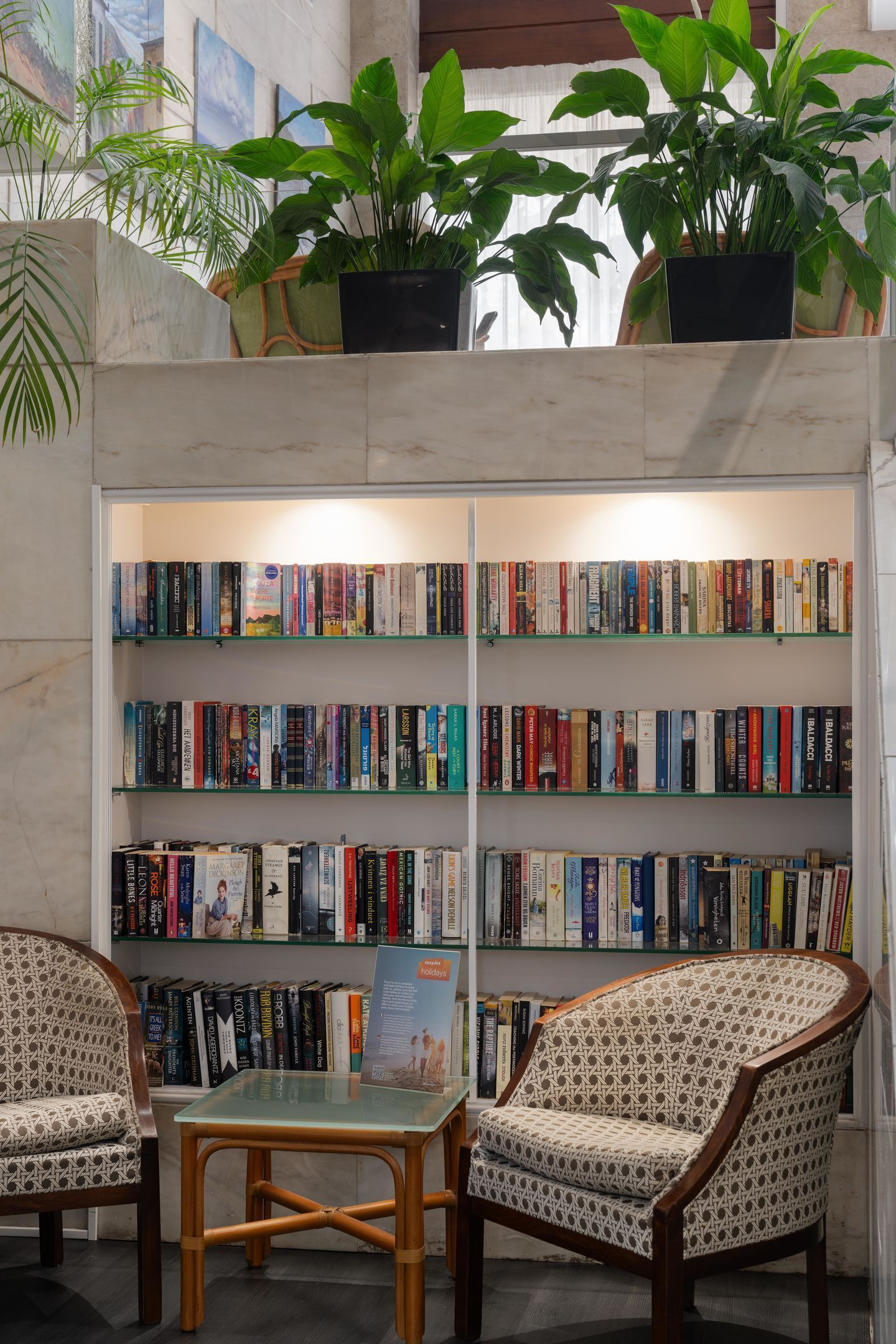 A library with bookshelves, two patterned chairs, a small table, and potted plants.
