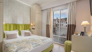 Une chambre d’hôtel avec un grand lit, une tête de lit recouverte d’un tissu vert, un balcon donnant sur des immeubles et un bureau avec une lampe.