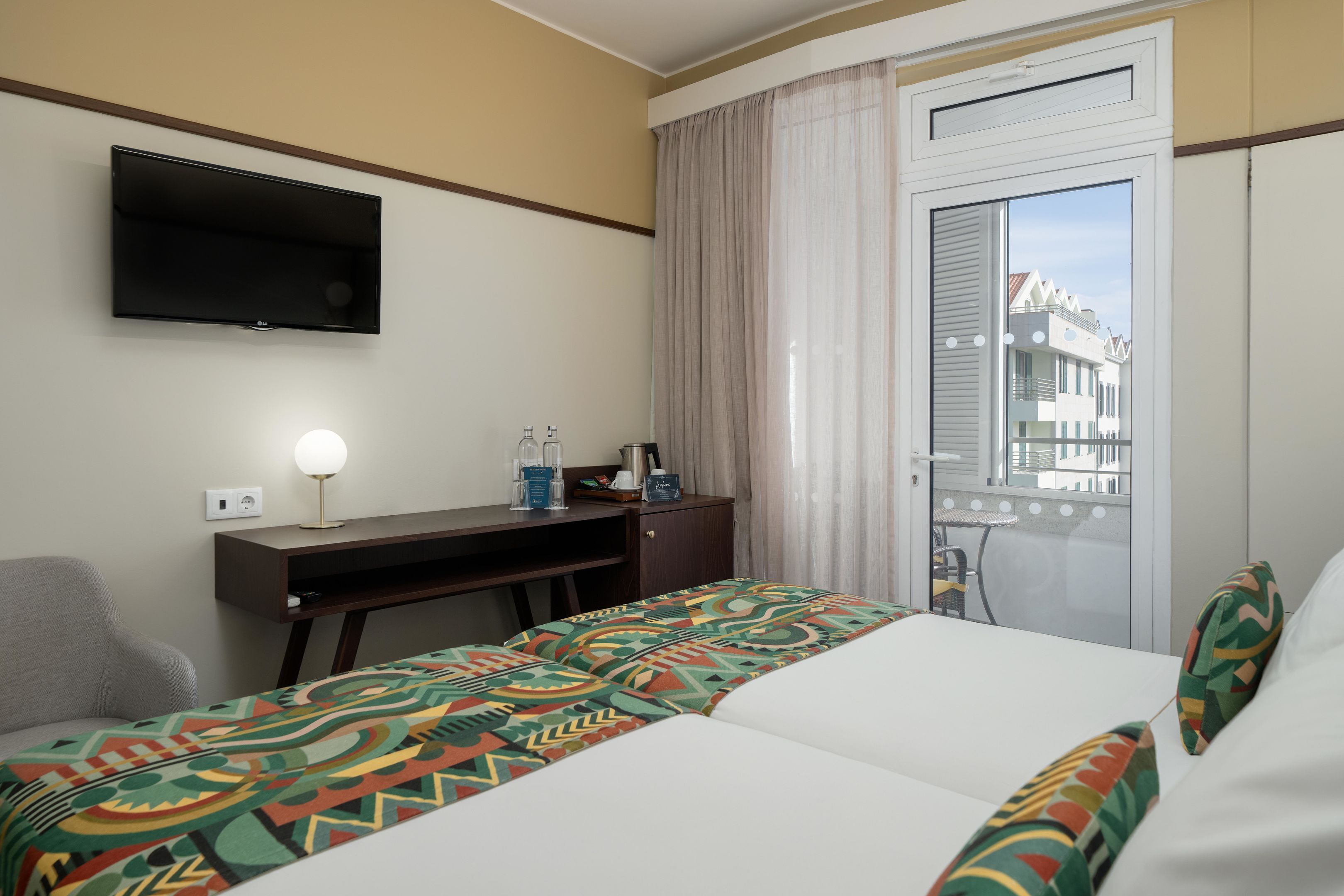 A hotel room with a bed, a desk with a lamp, a minibar, and a view of buildings through a window.