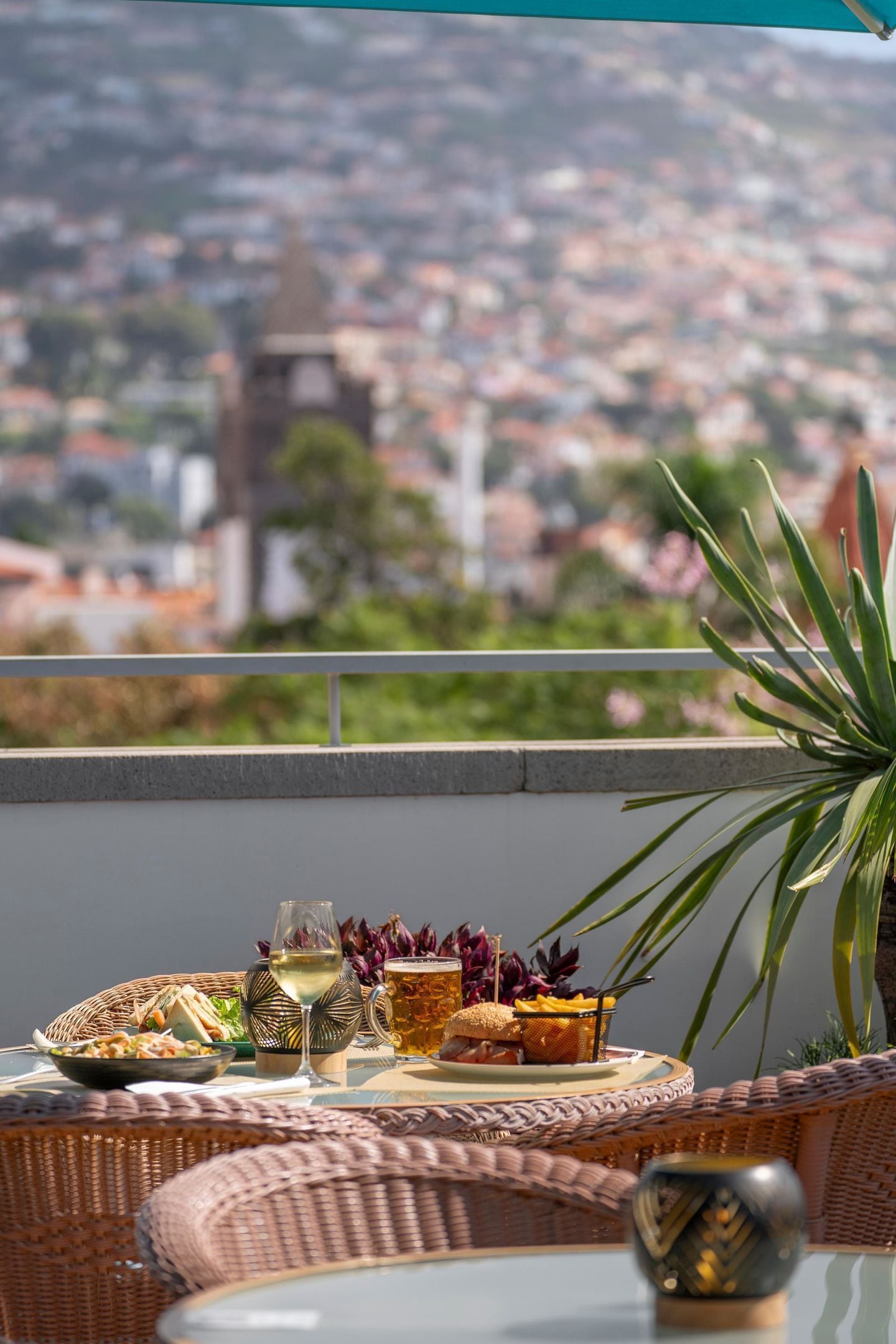 Un balcon agrémenté de meubles en osier, une table dressée avec de la nourriture et des boissons, et une vue sur un paysage urbain avec un beffroi.