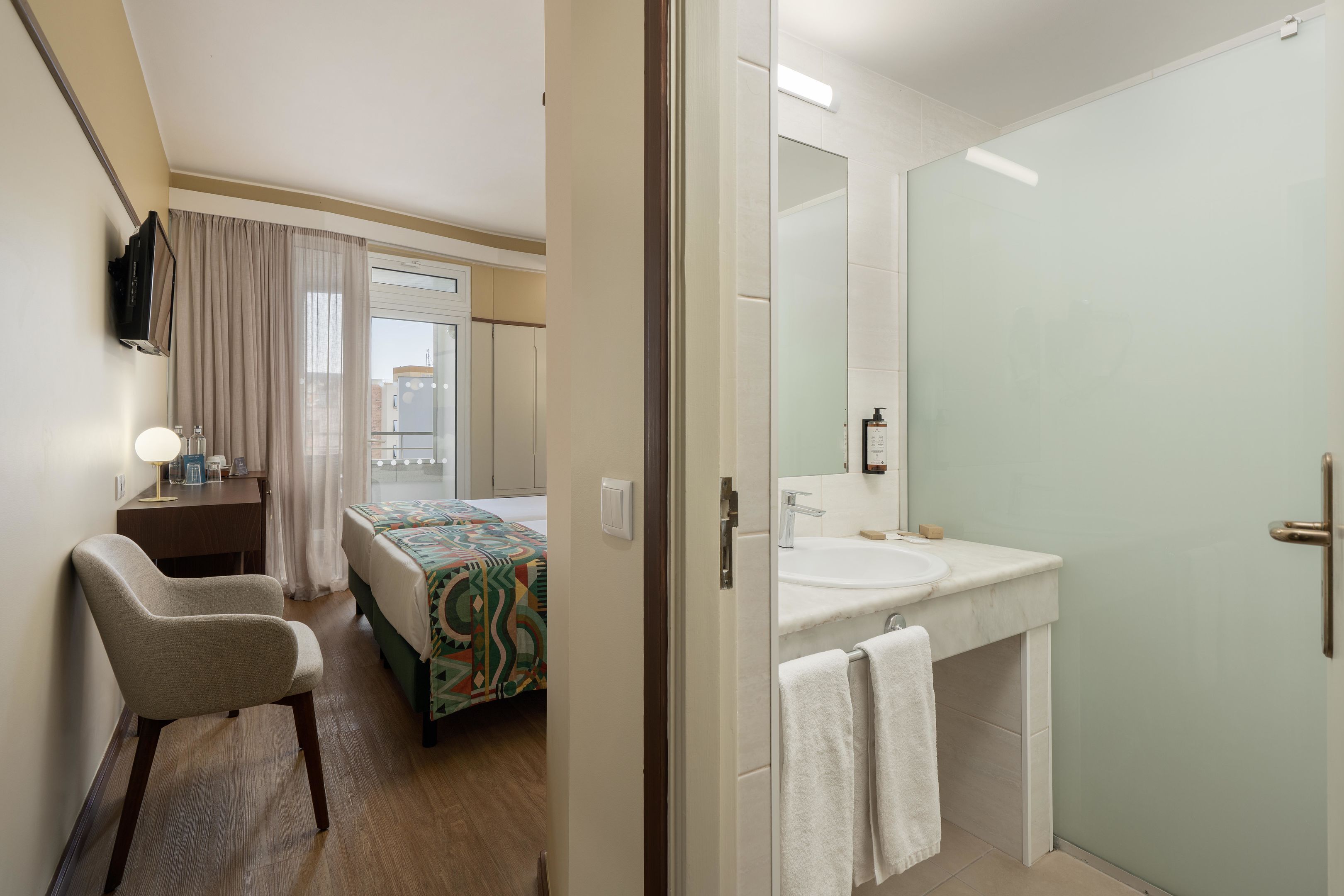 A hotel room with a bed, a desk, a chair, a bathroom with a sink and mirror, and a view of the city through the window.