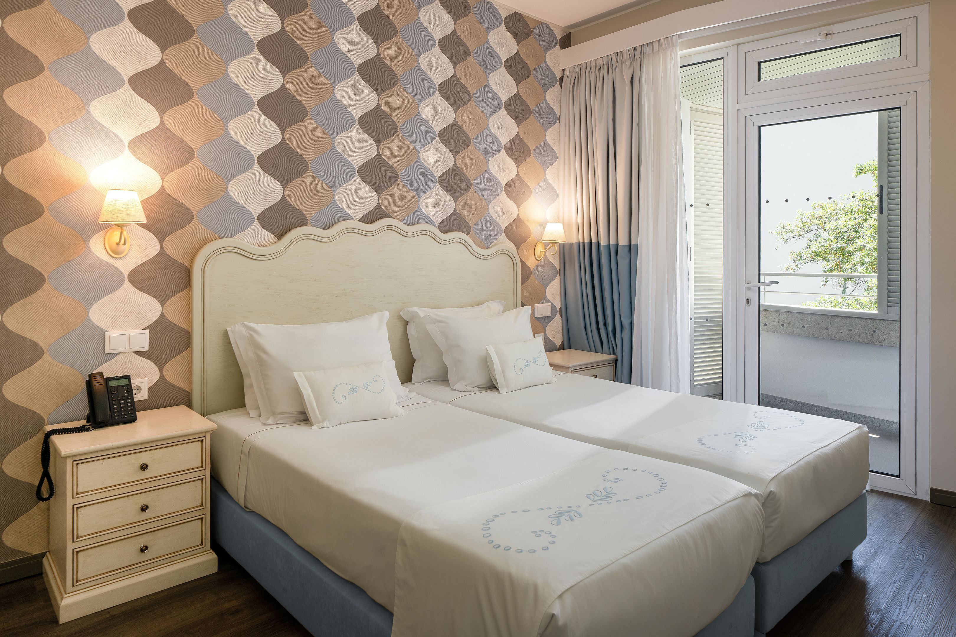Un lit double avec une literie blanche et une tête de lit décorative, une table de chevet avec une lampe et un mur orné d’un motif.