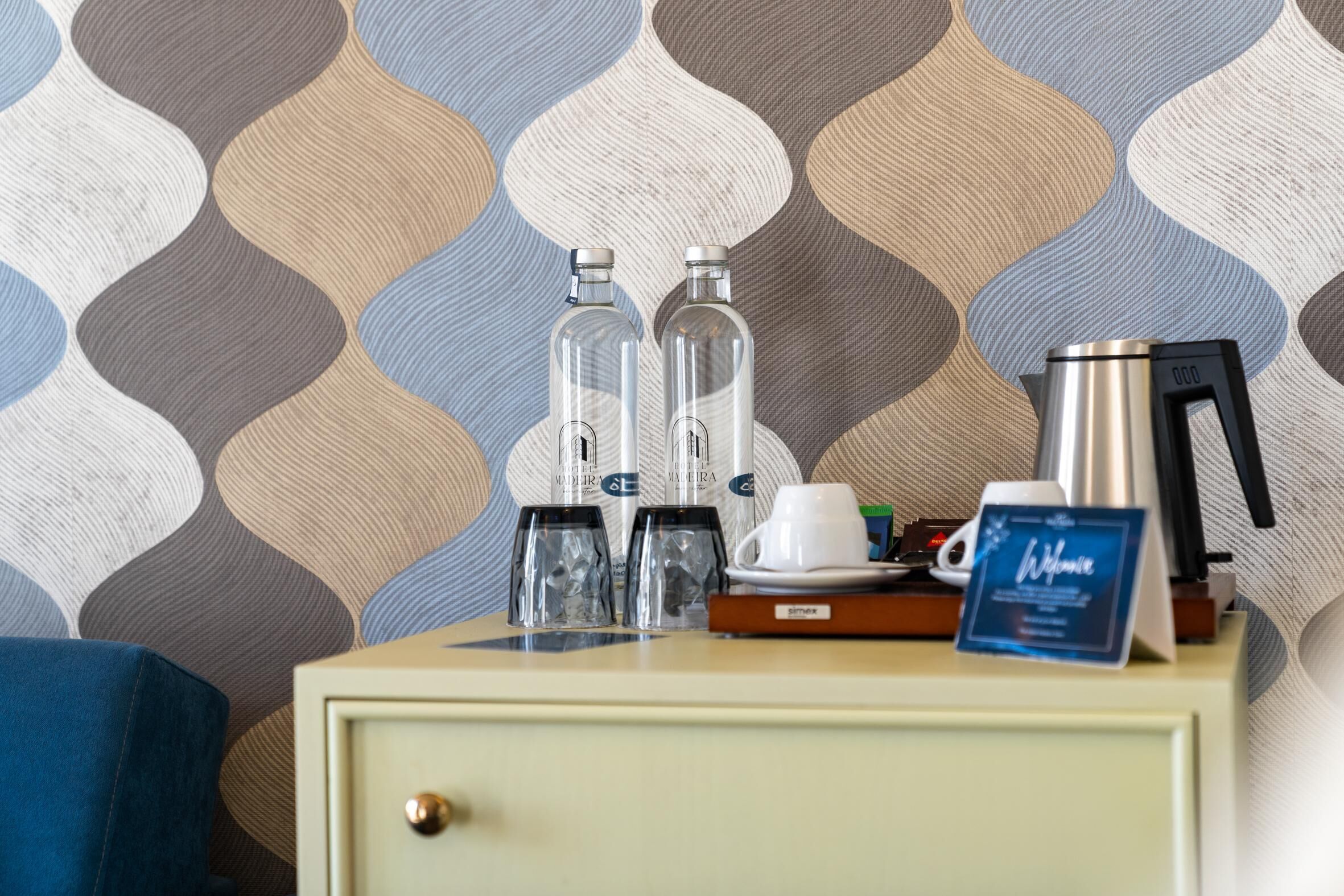 A minibar with two bottles, a coffee maker, a cup, and a sign on a wooden table against a patterned wallpaper.
