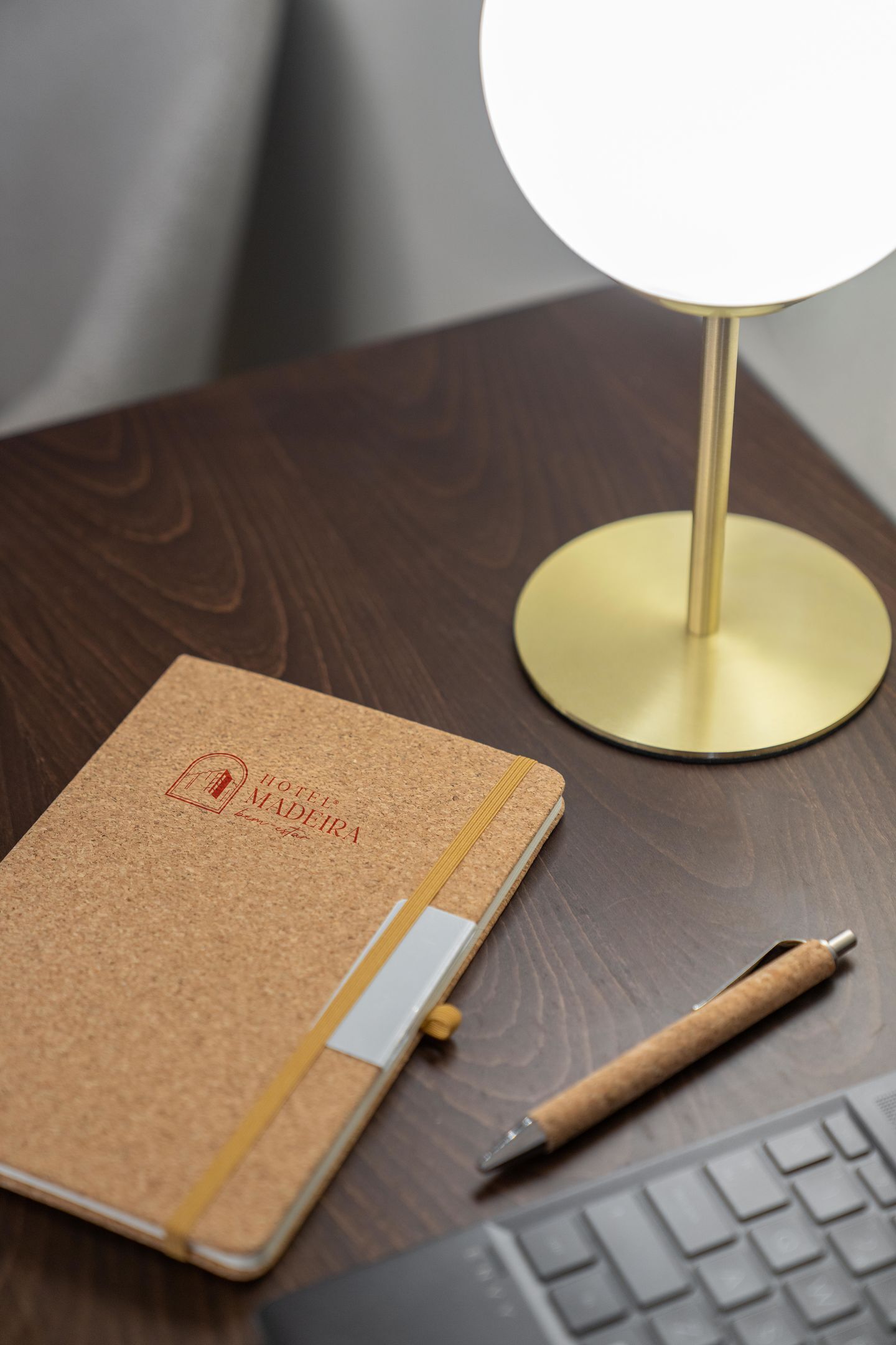 Un cuaderno de corcho con el logotipo de un hotel, un bolígrafo y una lámpara de escritorio sobre una superficie de madera.