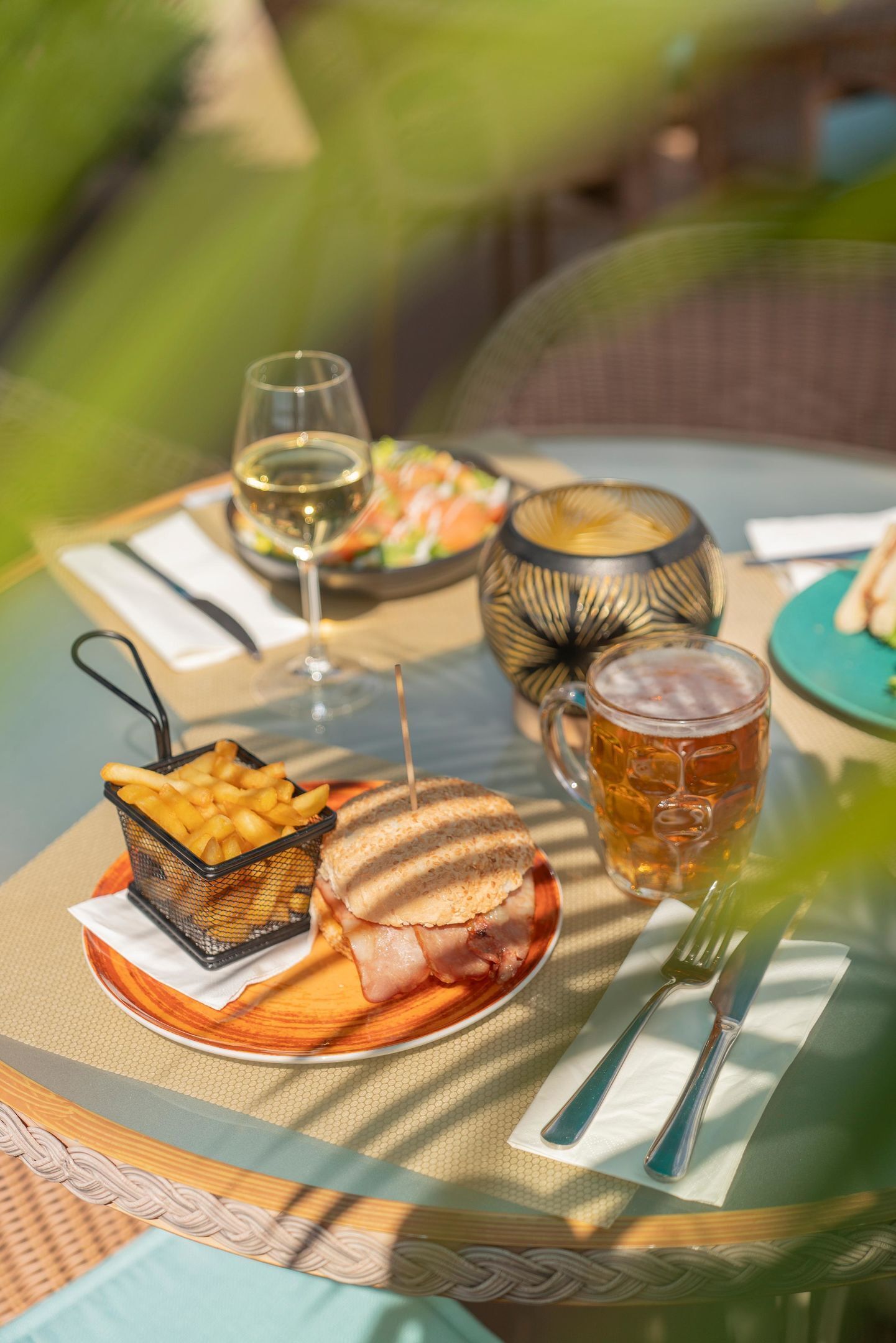 A table set with a sandwich, a glass of beer, a basket of fries, a glass of wine, and a decorative bowl.