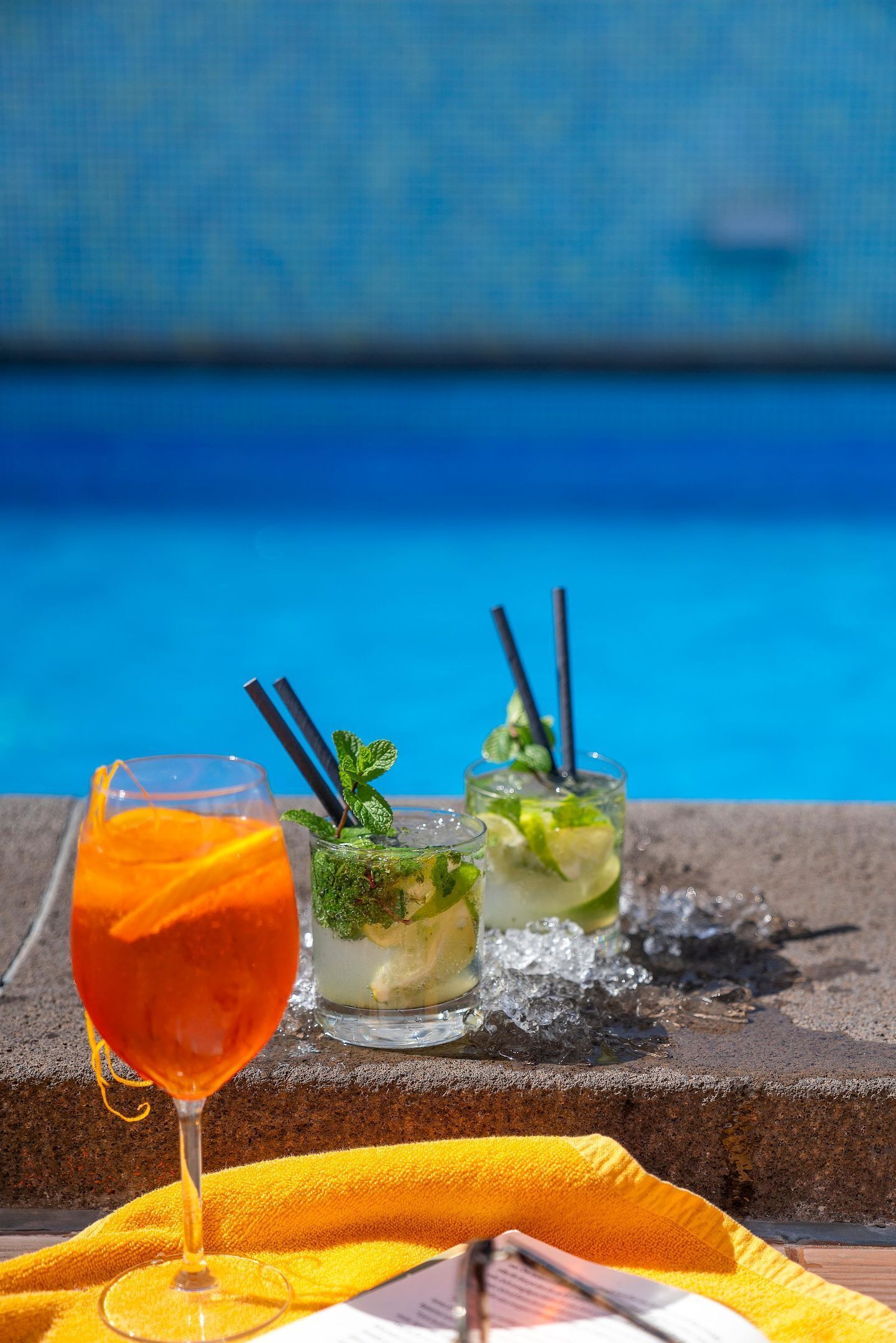 Un vaso de zumo de naranja, dos cócteles con pajitas y una servilleta amarilla junto a una piscina.