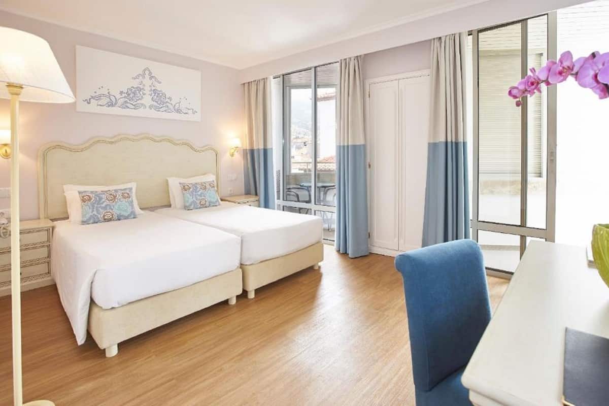 Ein Hotelzimmer mit Doppelbett, einem kleinen Tisch, einem Stuhl und einem großen Fenster mit Vorhängen.