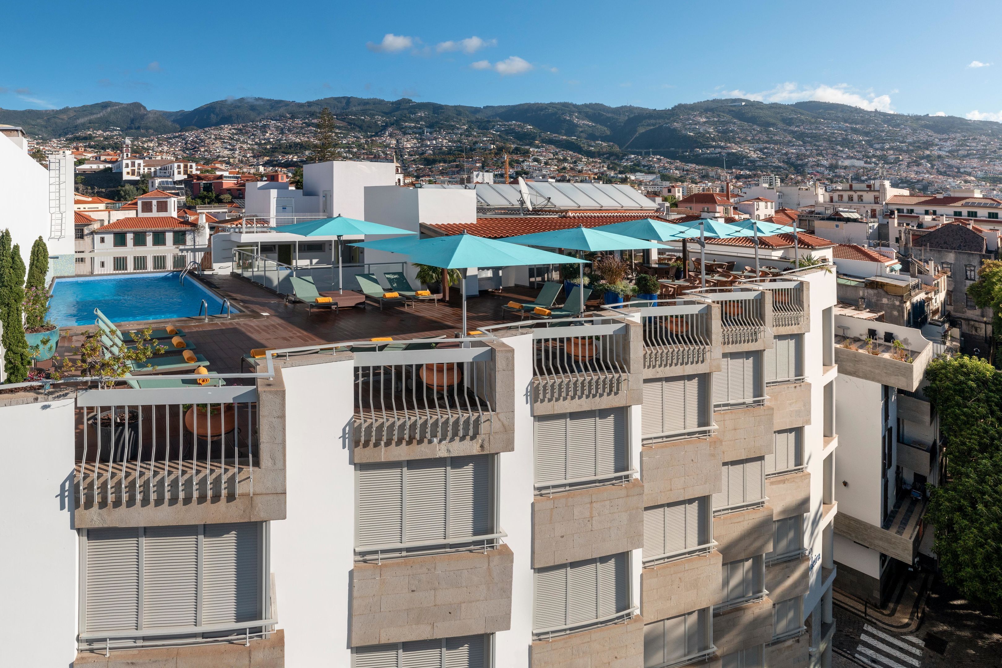 Un espace piscine aménagé sur le toit, doté de plusieurs chaises longues et de parasols, offrant une vue sur le paysage urbain avec des montagnes en arrière-plan.
