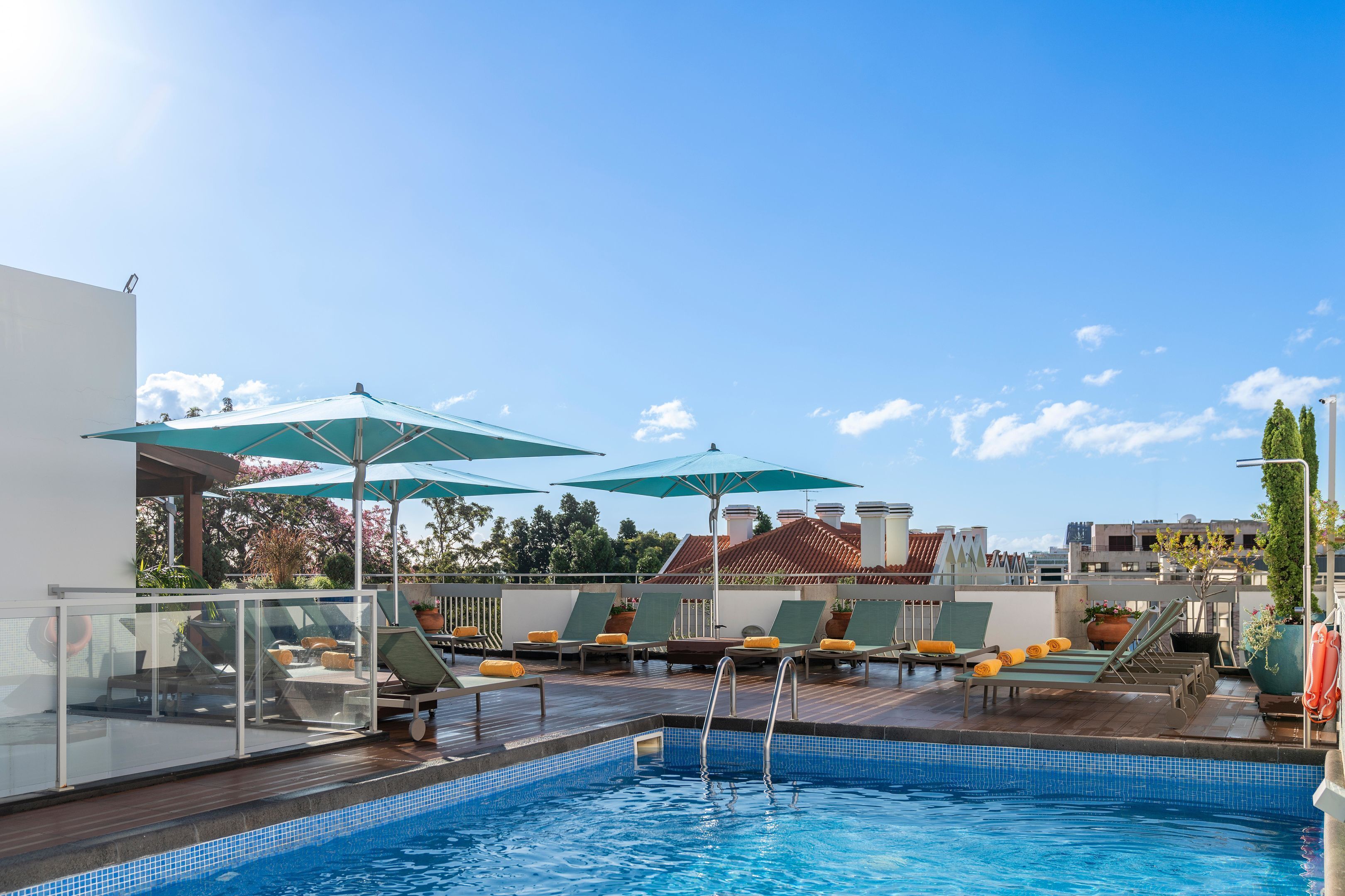 A rooftop pool area with lounge chairs and umbrellas, surrounded by a glass railing and a view of buildings in the background.