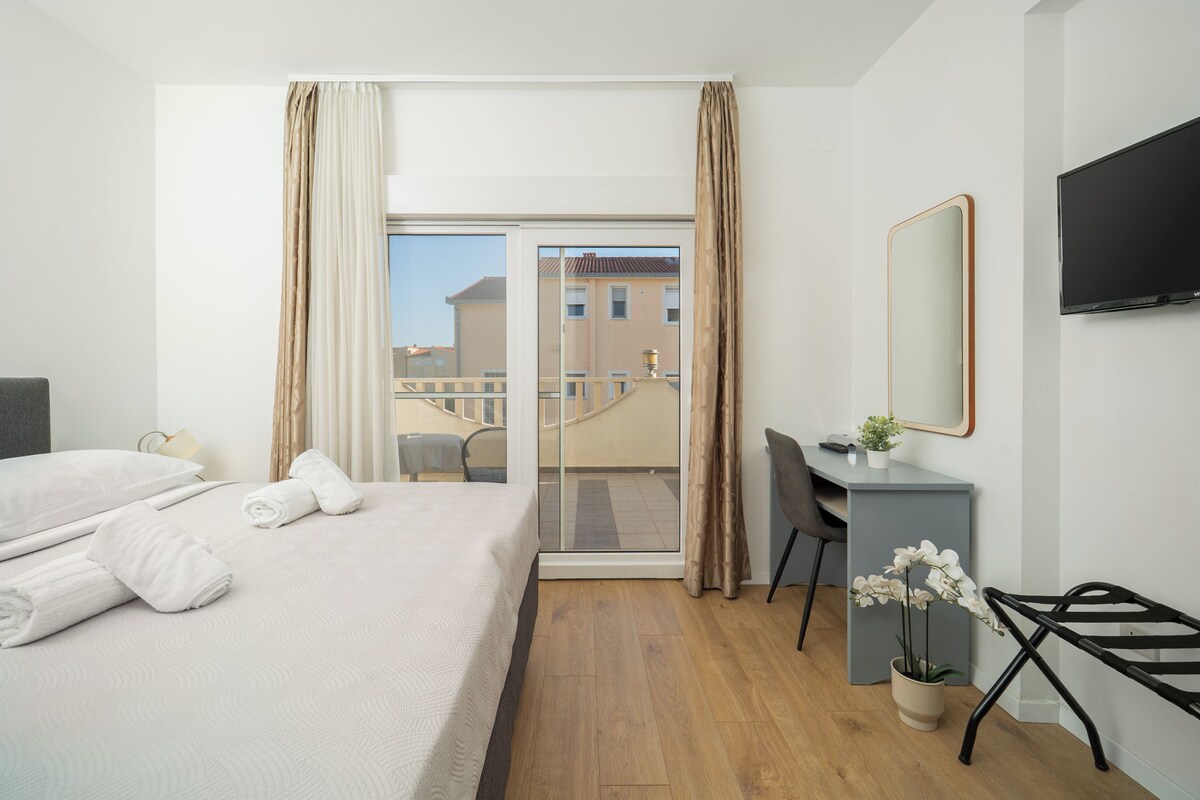 A hotel room with a bed, a desk, a chair, a TV, and a balcony with a view of buildings.