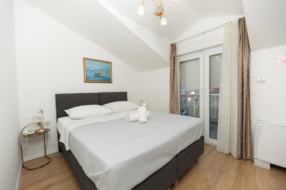 A bedroom with a bed, a bedside table with a lamp, a painting on the wall, and a view of the outside through a window.