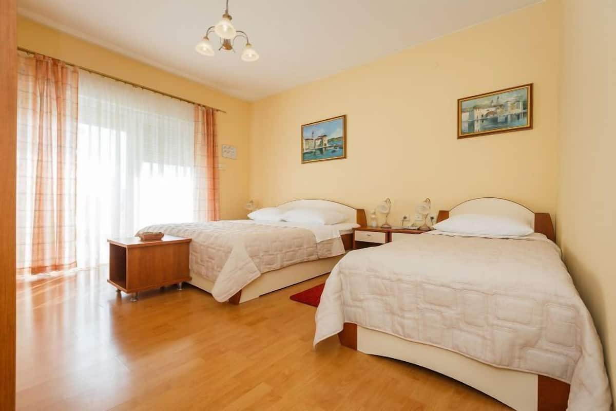 Quarto de hotel com duas camas, piso de madeira, mesa de cabeceira com abajur e janela com cortinas.