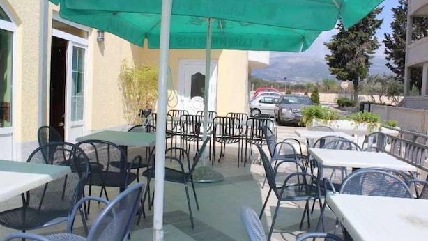 An outdoor cafe area with tables and chairs, a green umbrella, and a view of the street and mountains in the background. - Adria (Kastela)