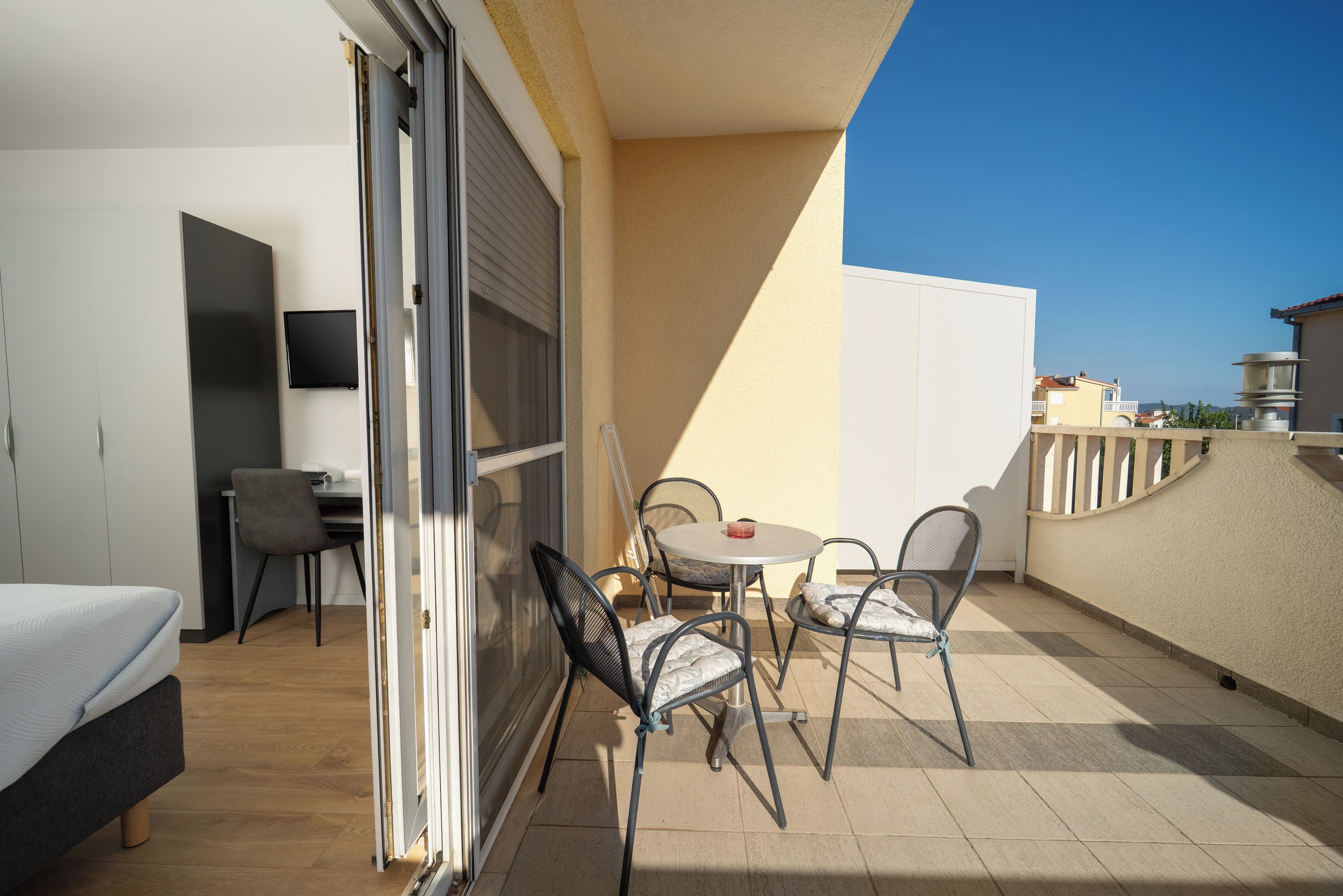 A balcony with a bed, a table, and chairs, a TV, and a view of the sea.