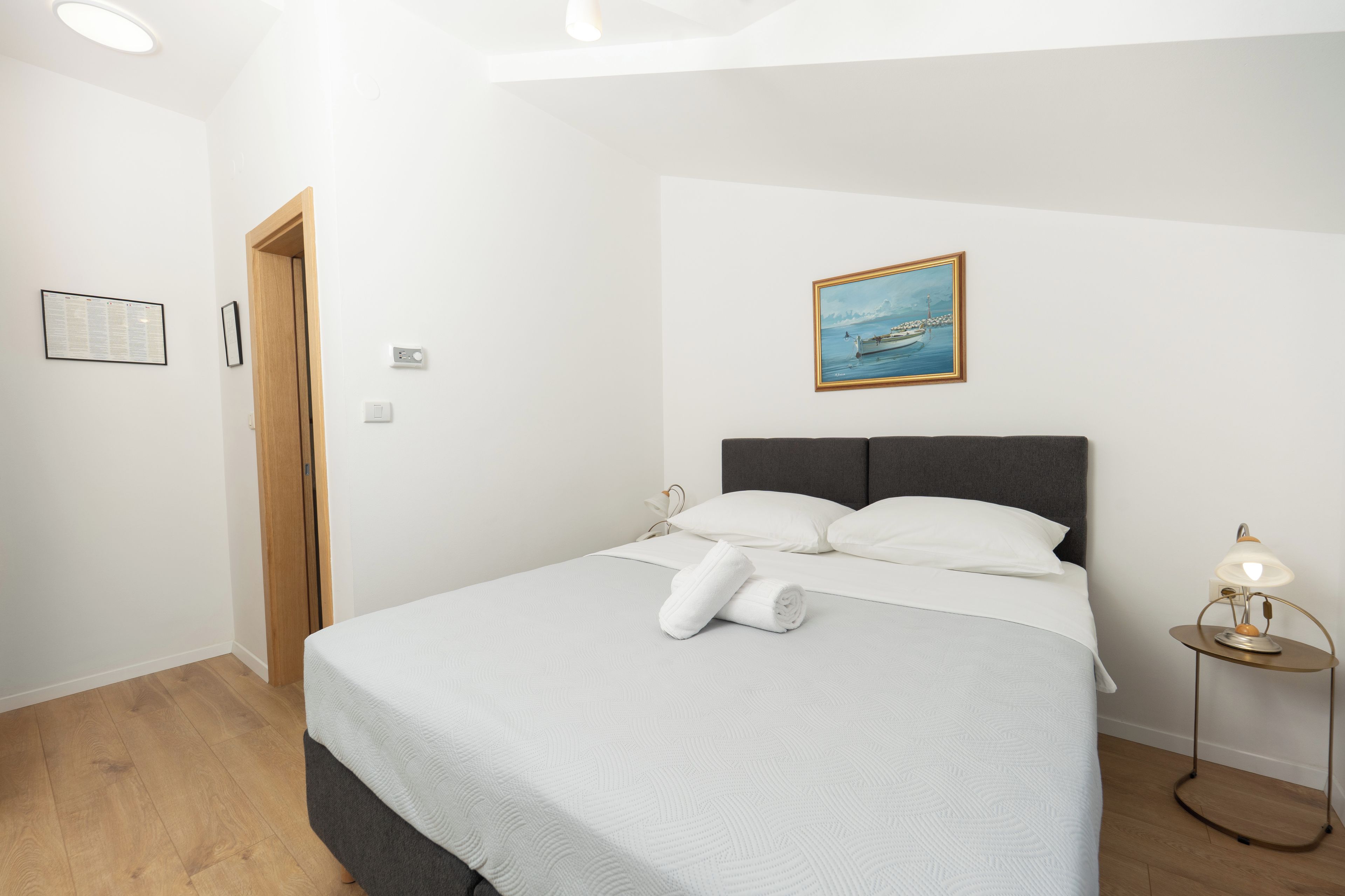 Ein ordentlich bezogenes Bett mit weißer Decke und Kissen, ein Bild von einem Boot an der Wand, ein kleiner Nachttisch mit einer Lampe und eine Holztür.