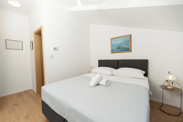 A neatly made bed with a white comforter and pillows, a painting of a boat on the wall, a small bedside table with a lamp, and a wooden door.