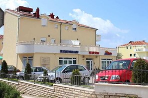Un edificio con restaurante y tienda en la planta baja, varios coches aparcados y un vehículo rojo en primer plano.