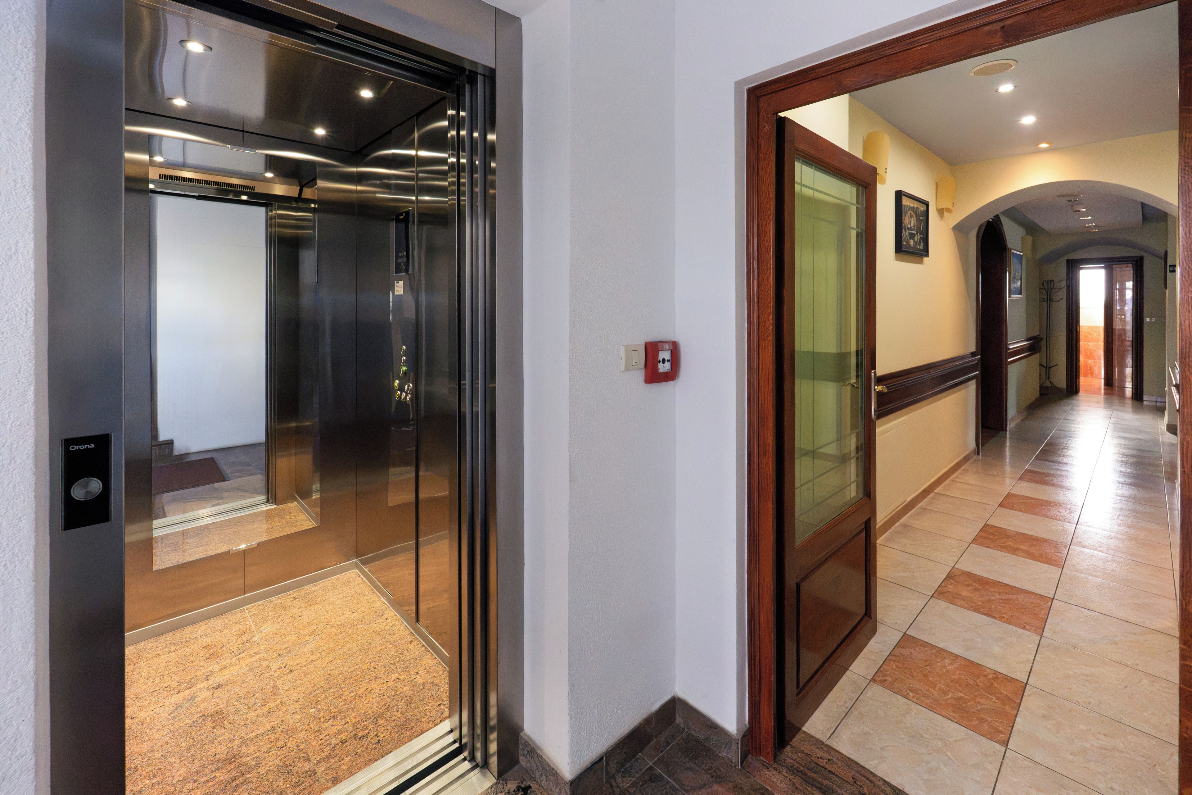Un ascensor modern amb porta de vidre, un passadís amb sòl de rajola i un marc de porta de fusta.