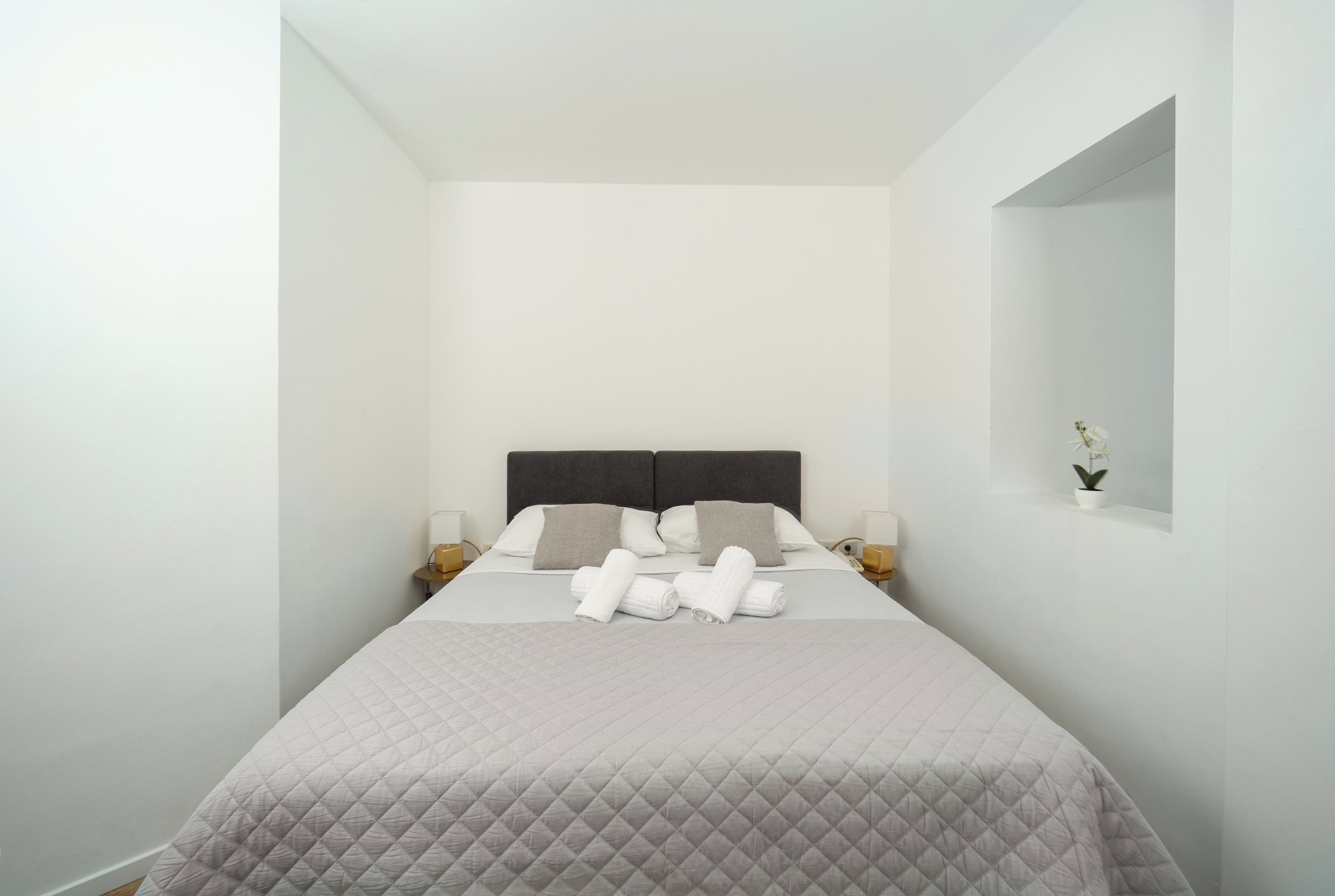 A hotel room with a bed, pillows, a nightstand, a lamp, and a small potted plant.