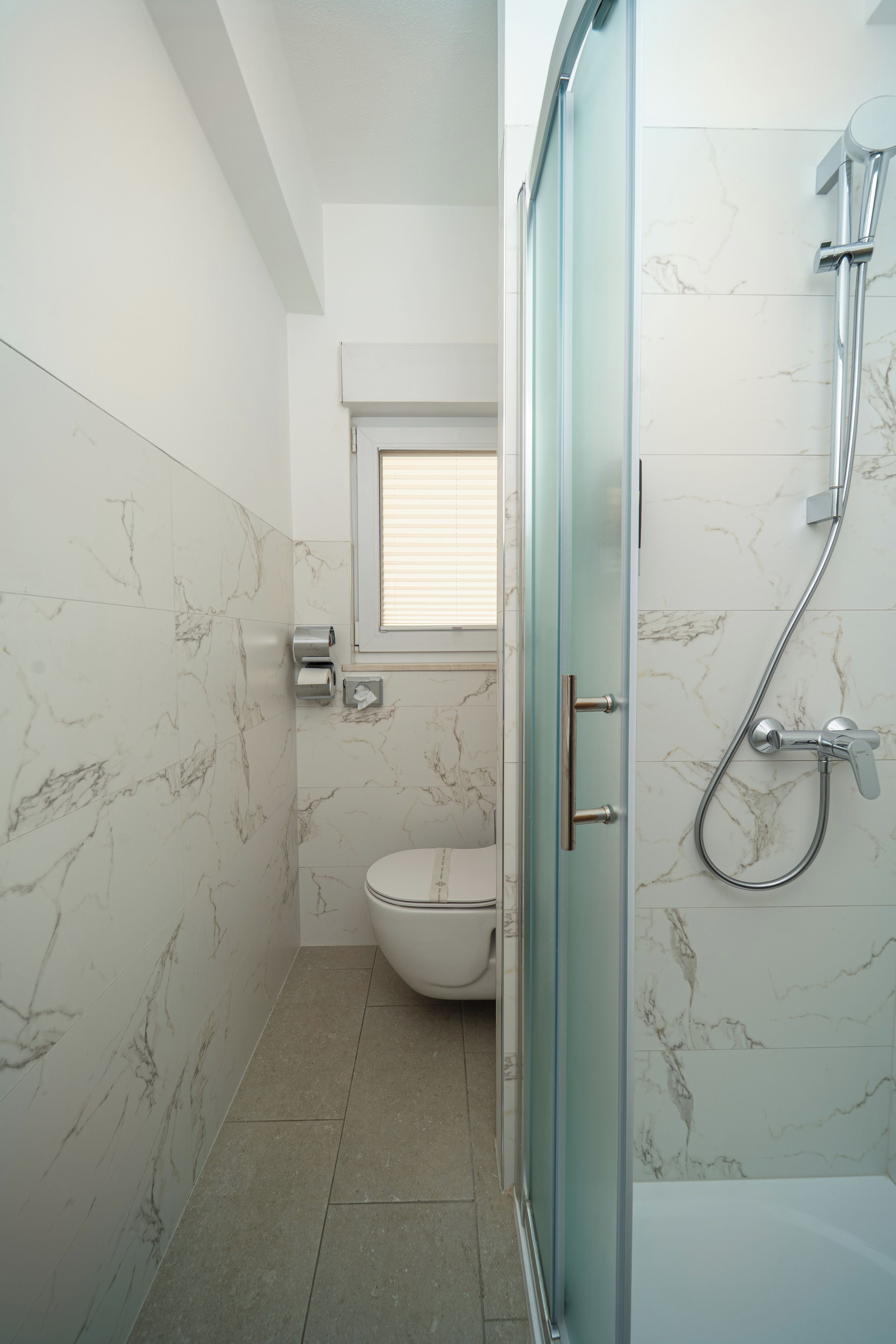Un baño con cabina de ducha de vidrio, inodoro y ventana con persianas.