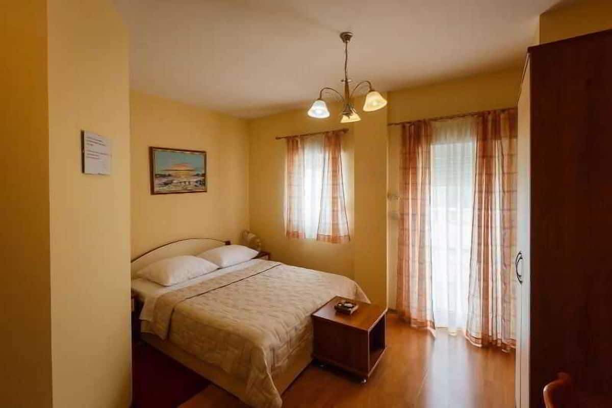 Quarto com cama, mesa de cabeceira, luminária pendente e janela com cortinas.