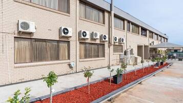 Edifício com várias unidades de ar condicionado, fachada de tijolos e área ajardinada com árvores pequenas.