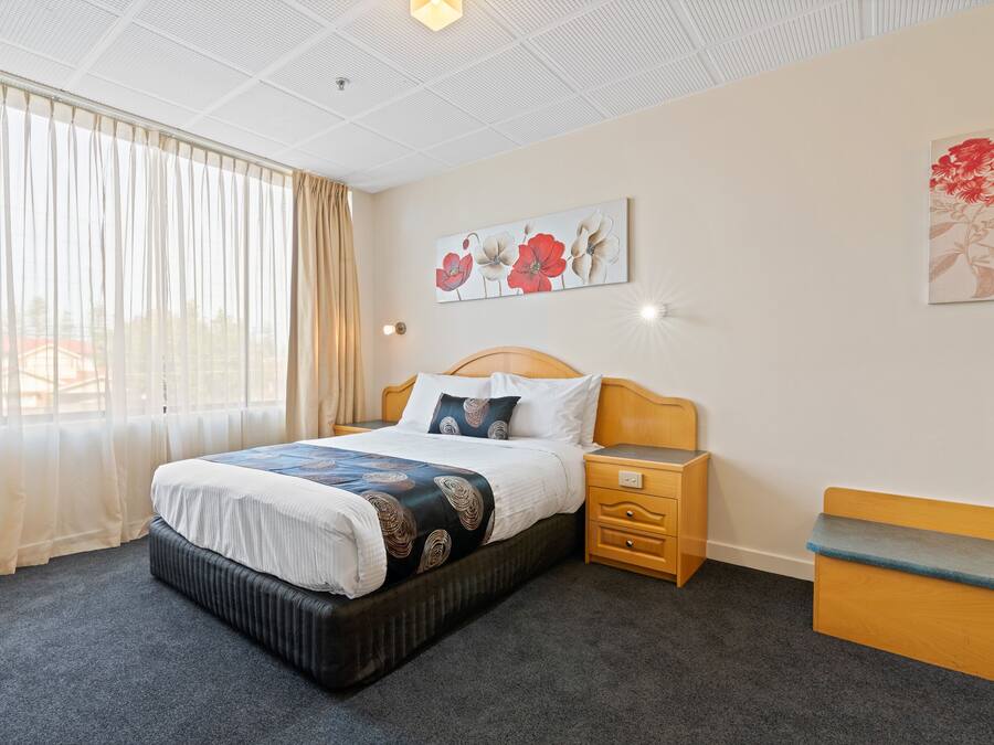 Quarto de hotel com cama, mesa de cabeceira e um pequeno banco de madeira.