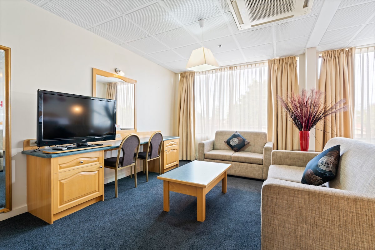 A hotel room with a TV, wooden desk, chairs, a sofa, and a coffee table.