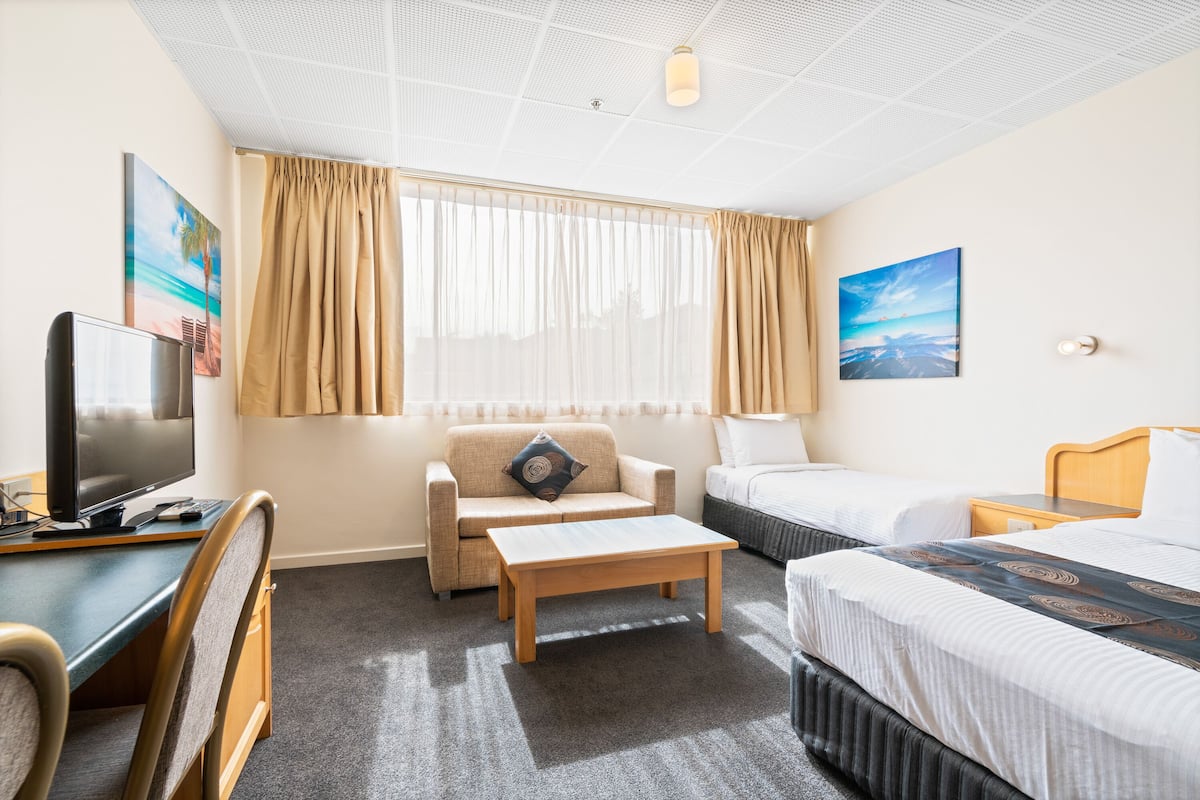 A hotel room with a bed, a sofa, a desk with a TV, a coffee table, and a window with curtains.