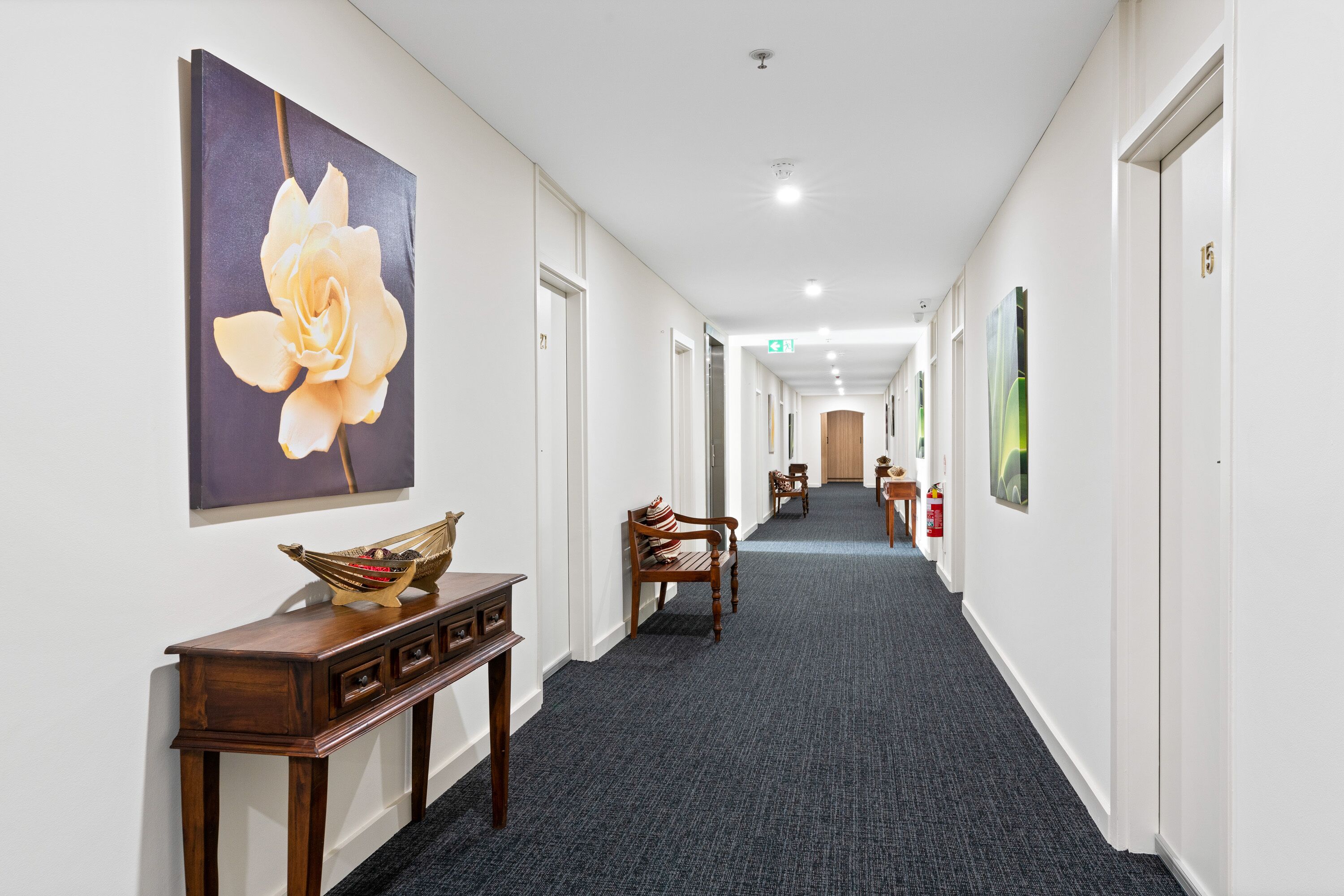 A hotel hallway with a long carpeted corridor, wooden furniture, and artwork on the walls.