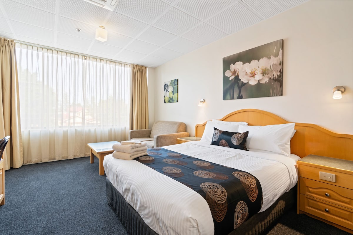 A hotel room with a large bed, a wooden nightstand, a chair, a desk, and a window with curtains.