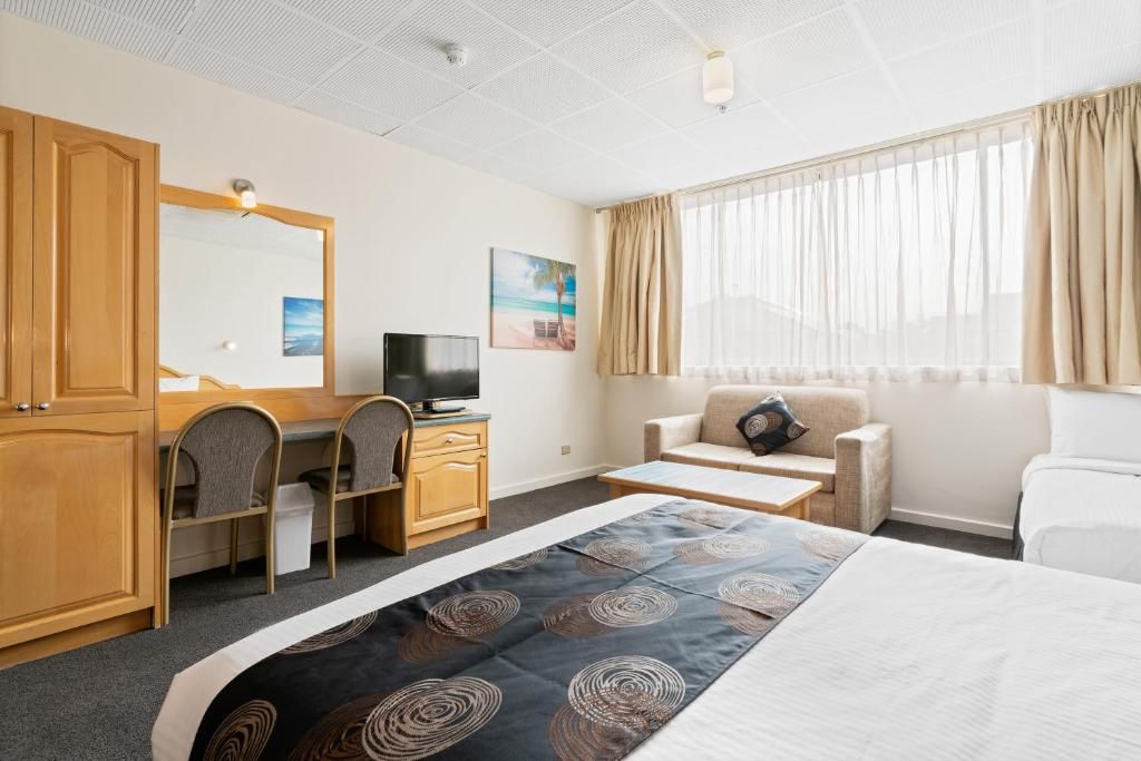 A hotel room with a bed, a desk with a computer, a television, a sofa, and a window with curtains.