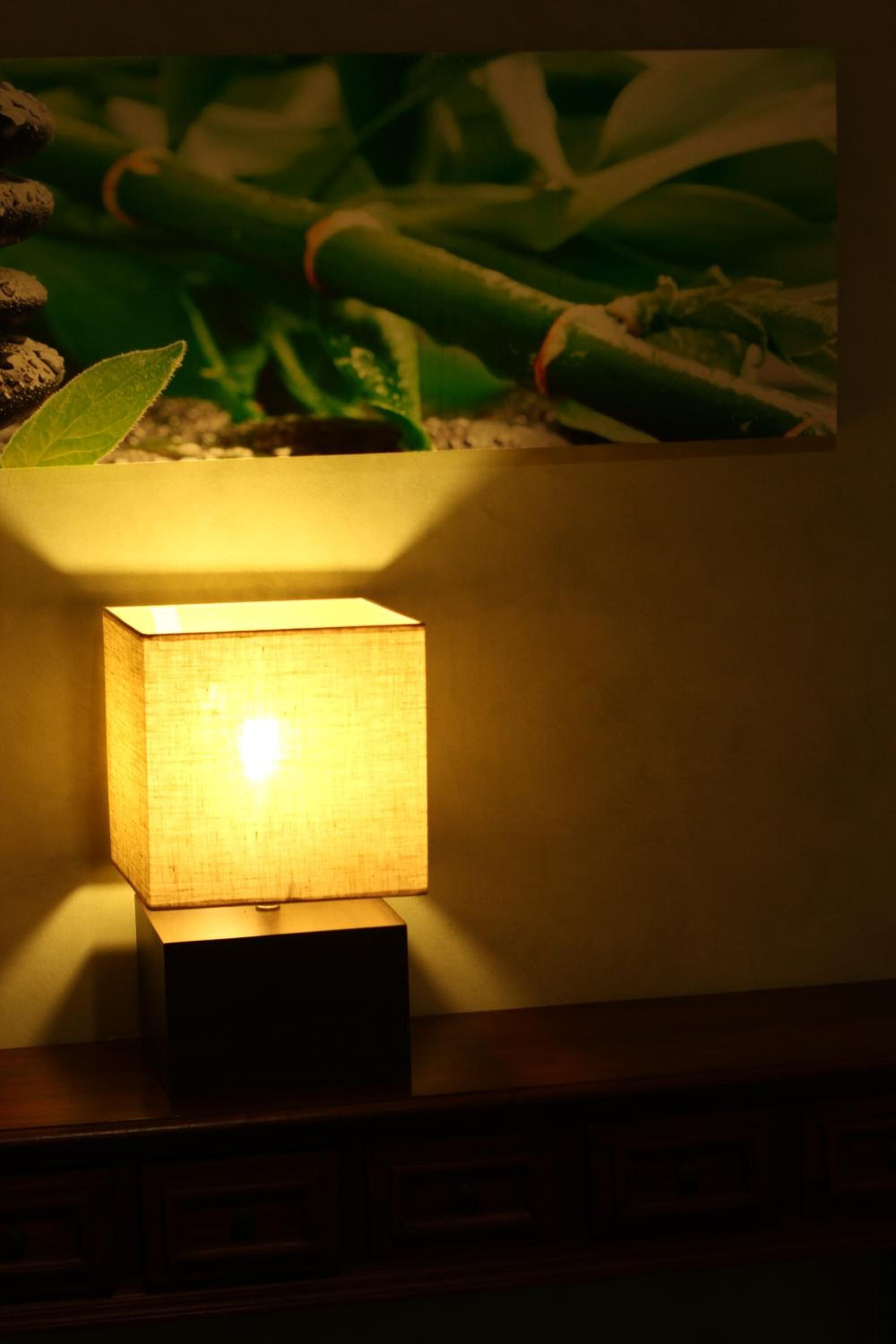 A square table lamp with a fabric shade, placed on a wooden surface against a wall with a bamboo plant backdrop.