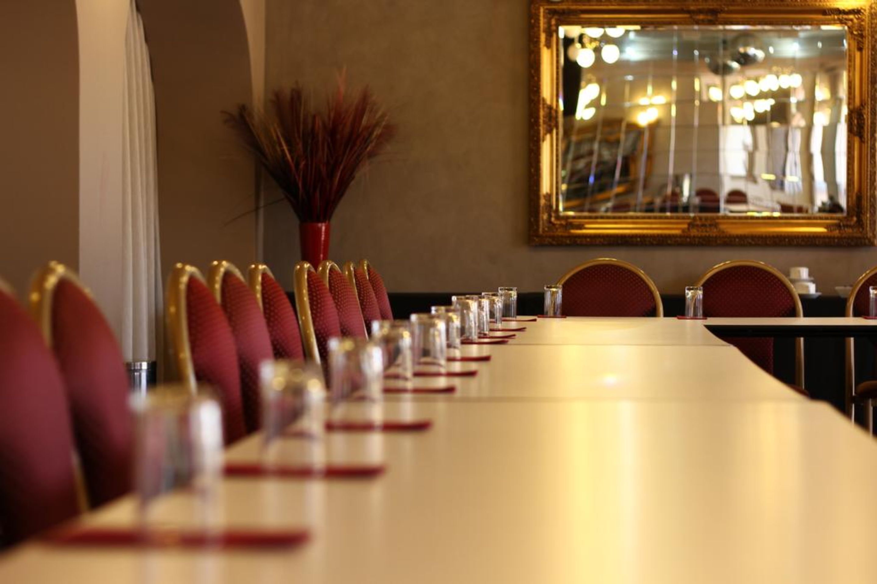 A long dining table with red chairs, a vase with dried plants, and a mirror on the wall.