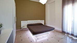 A bedroom with a bed, a bedside table, a nightstand, a lamp, a window with curtains, and a wall-mounted light fixture. - Hyencos Hotel Calos (Ugento)
