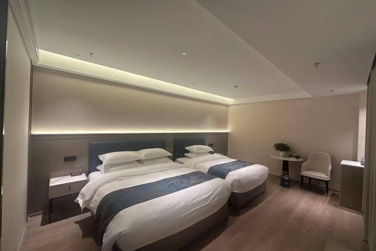 A modern hotel room with two beds, a bedside table, a chair, and a small table with a plant.