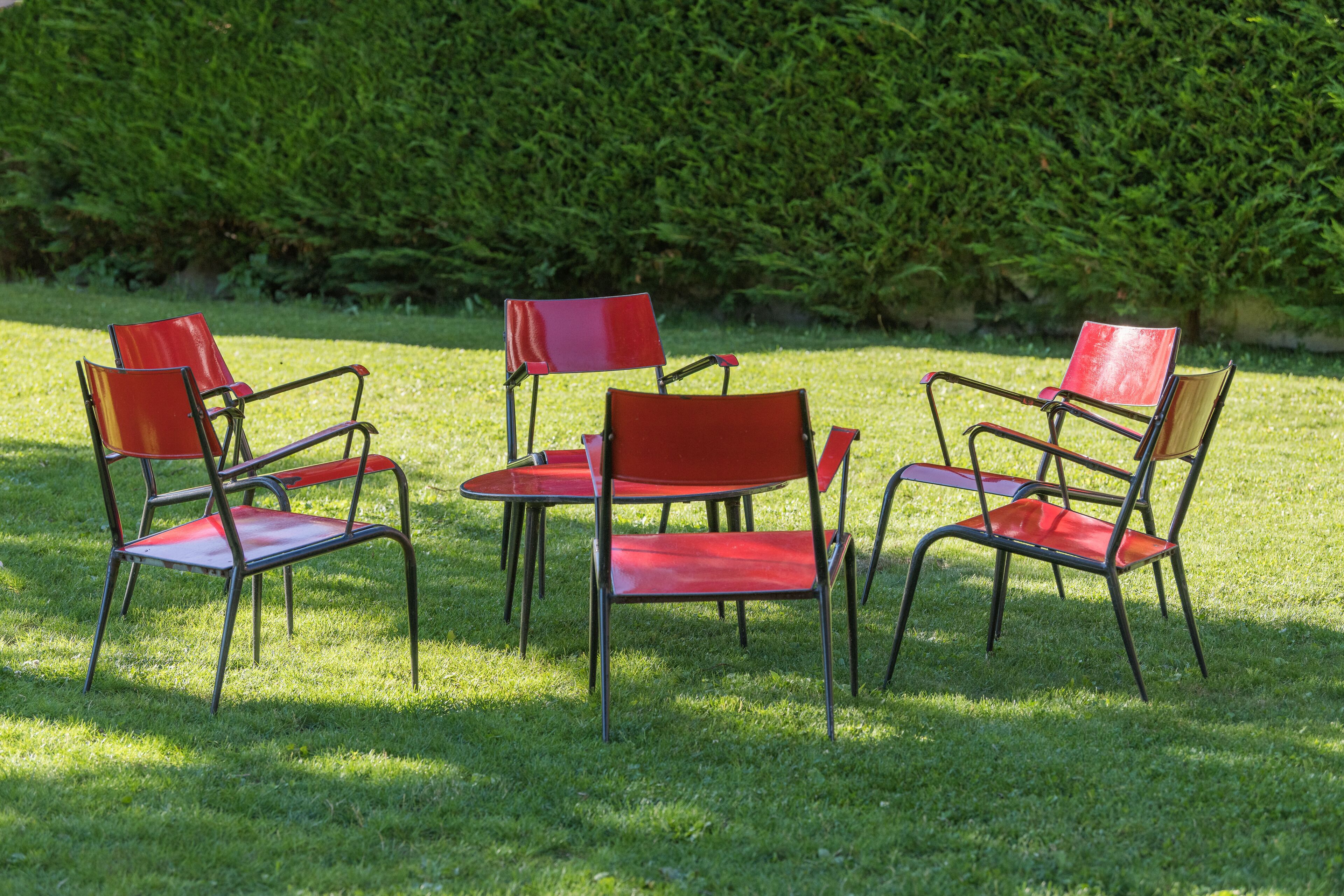 A set of four red chairs arranged around a small table on a grassy lawn with a hedge in the background.
