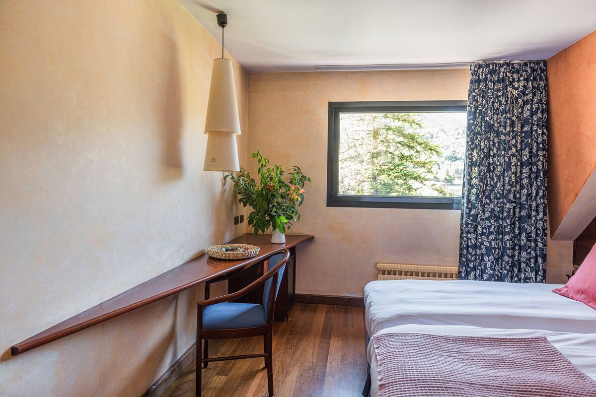 A bedroom with a bed, a desk with a chair, a potted plant, and a window with curtains.