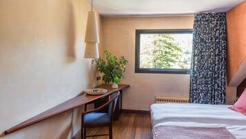 A bedroom with a bed, a desk with a chair, a potted plant, and a window with curtains.
