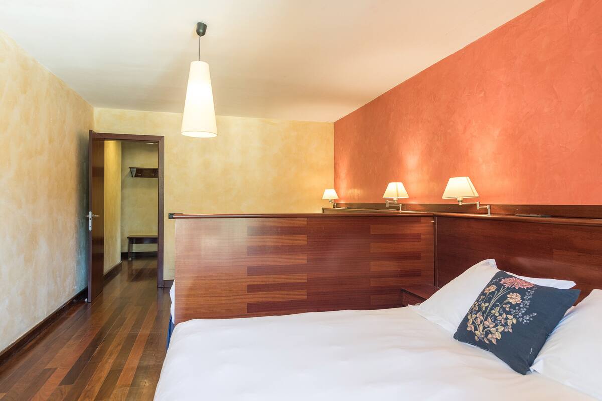 A hotel room with a wooden headboard, a bed with white linens, two bedside lamps, and a door leading to a hallway.